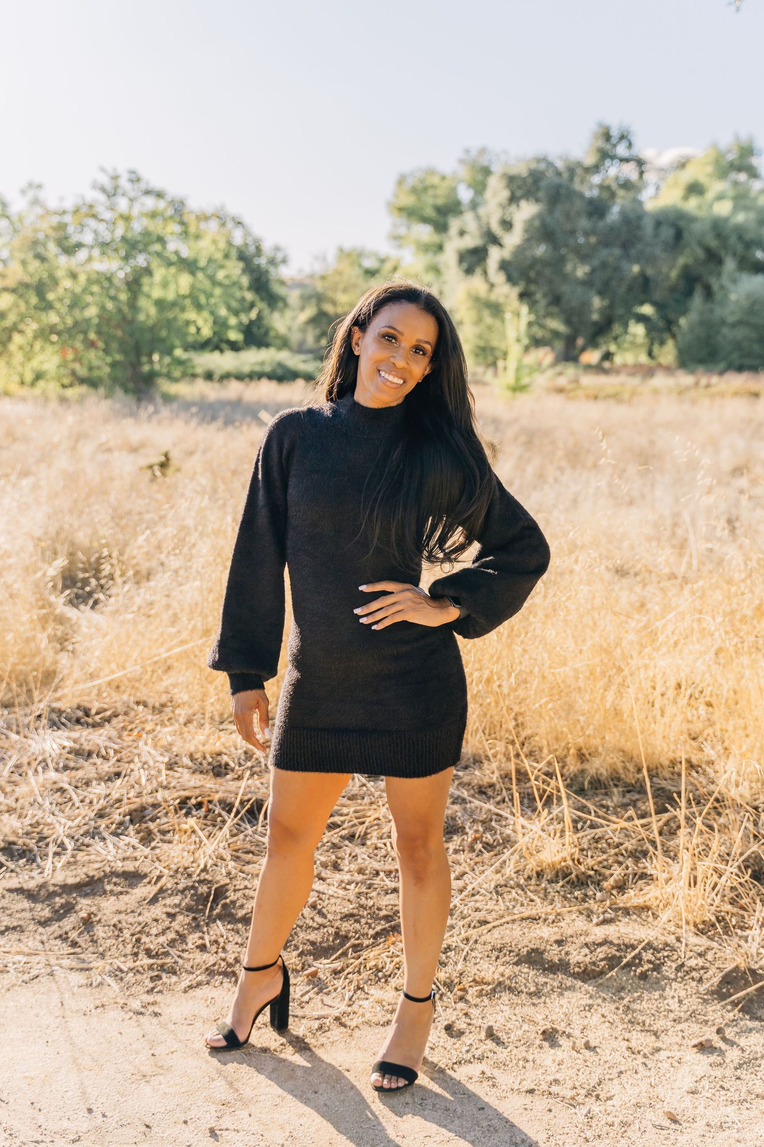 A person in a black sweater dress and heels poses confidently in a sunlit field with dried grass and trees.
