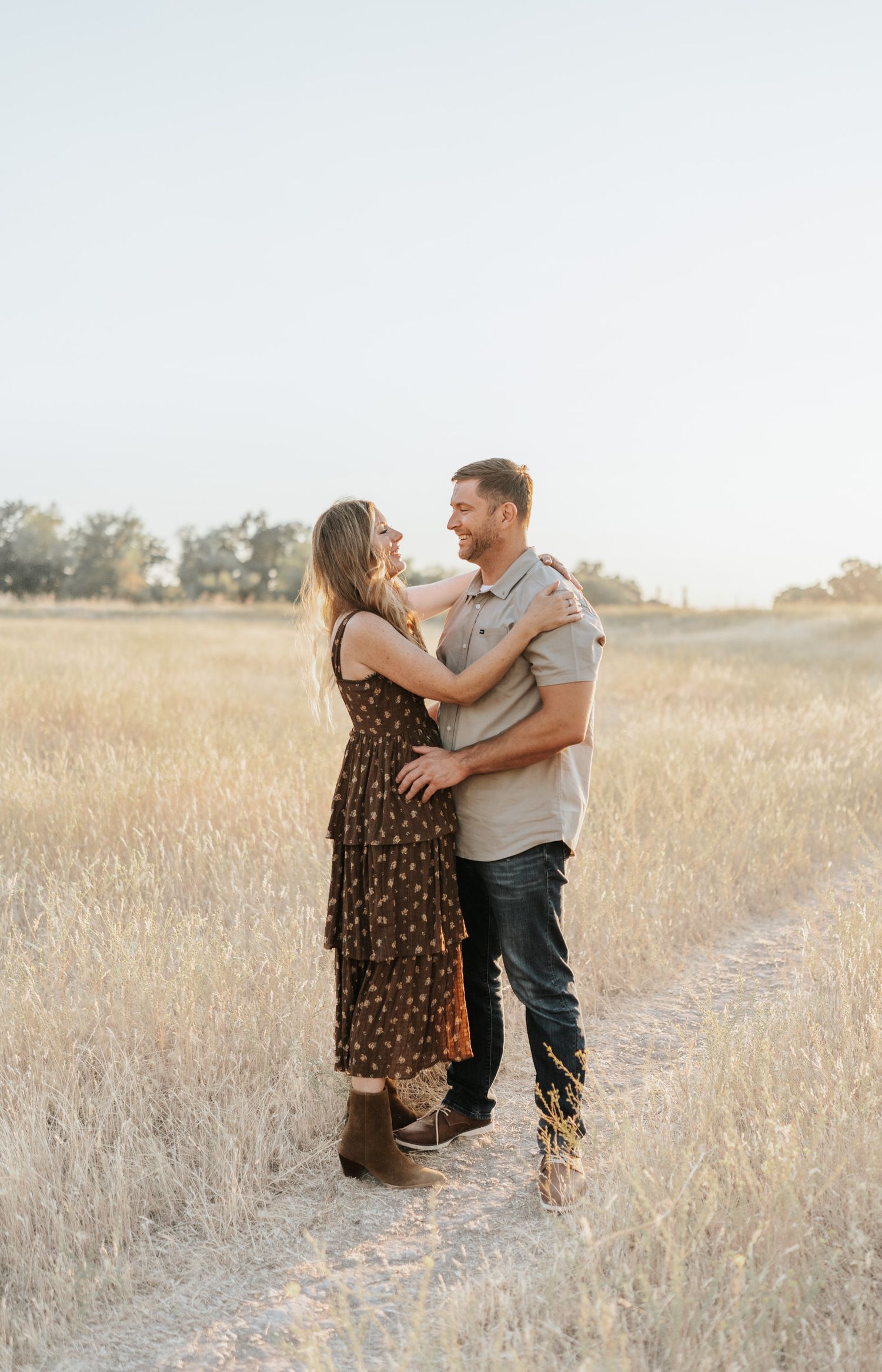 A couple embraces lovingly while standing in a golden sunlit field during a romantic outdoor photoshoot.