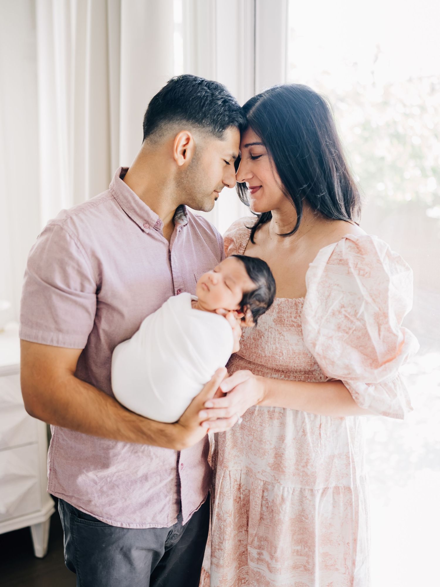 A couple tenderly touches foreheads while holding their swaddled newborn baby in a bright, airy room.