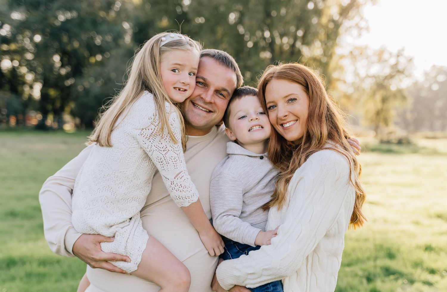 Happy family of four smiling outdoors in a sunny green park, parents holding two young children in white sweaters.