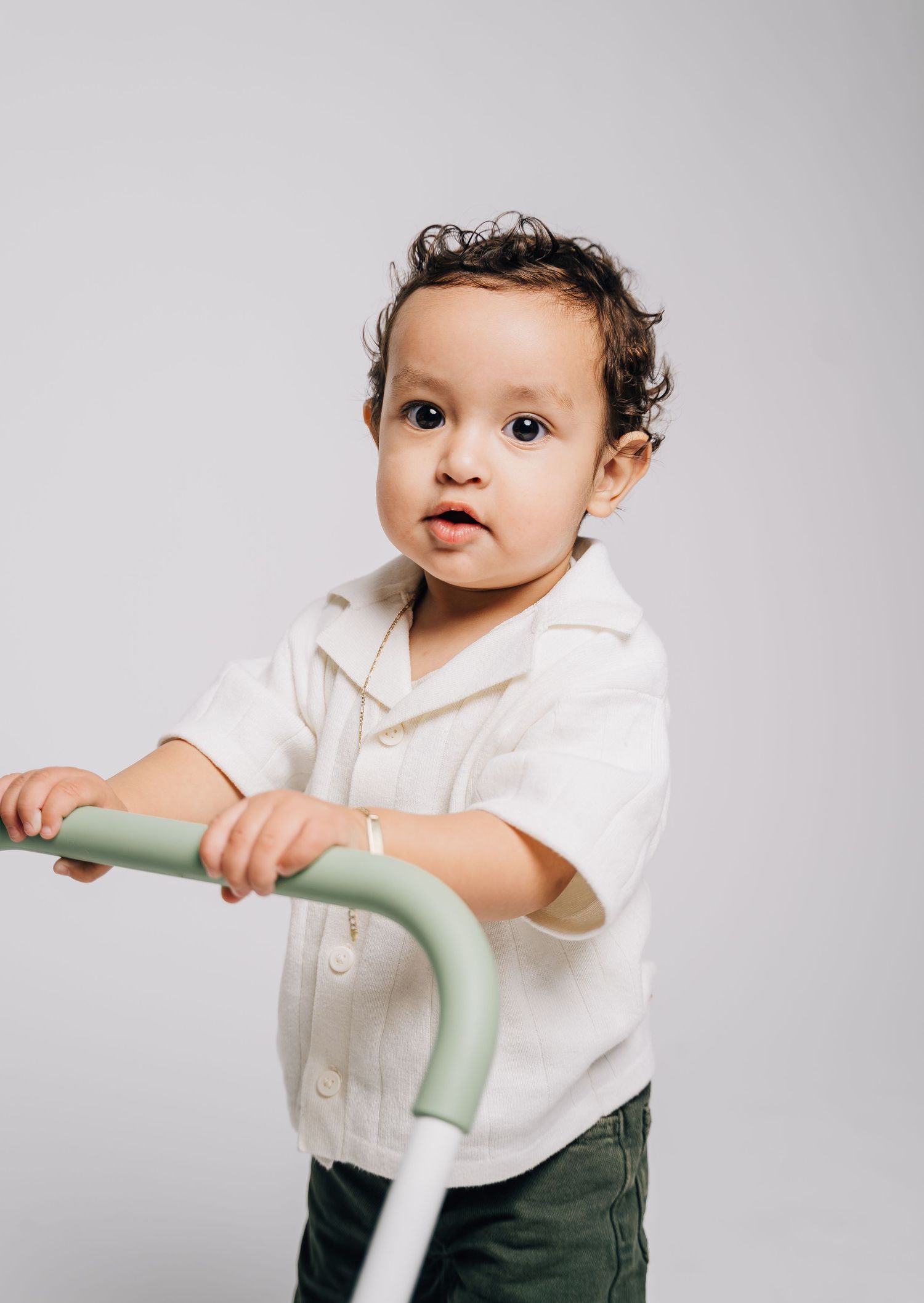 Cute toddler in white shirt holding a sage green baby walker against a light gray background.