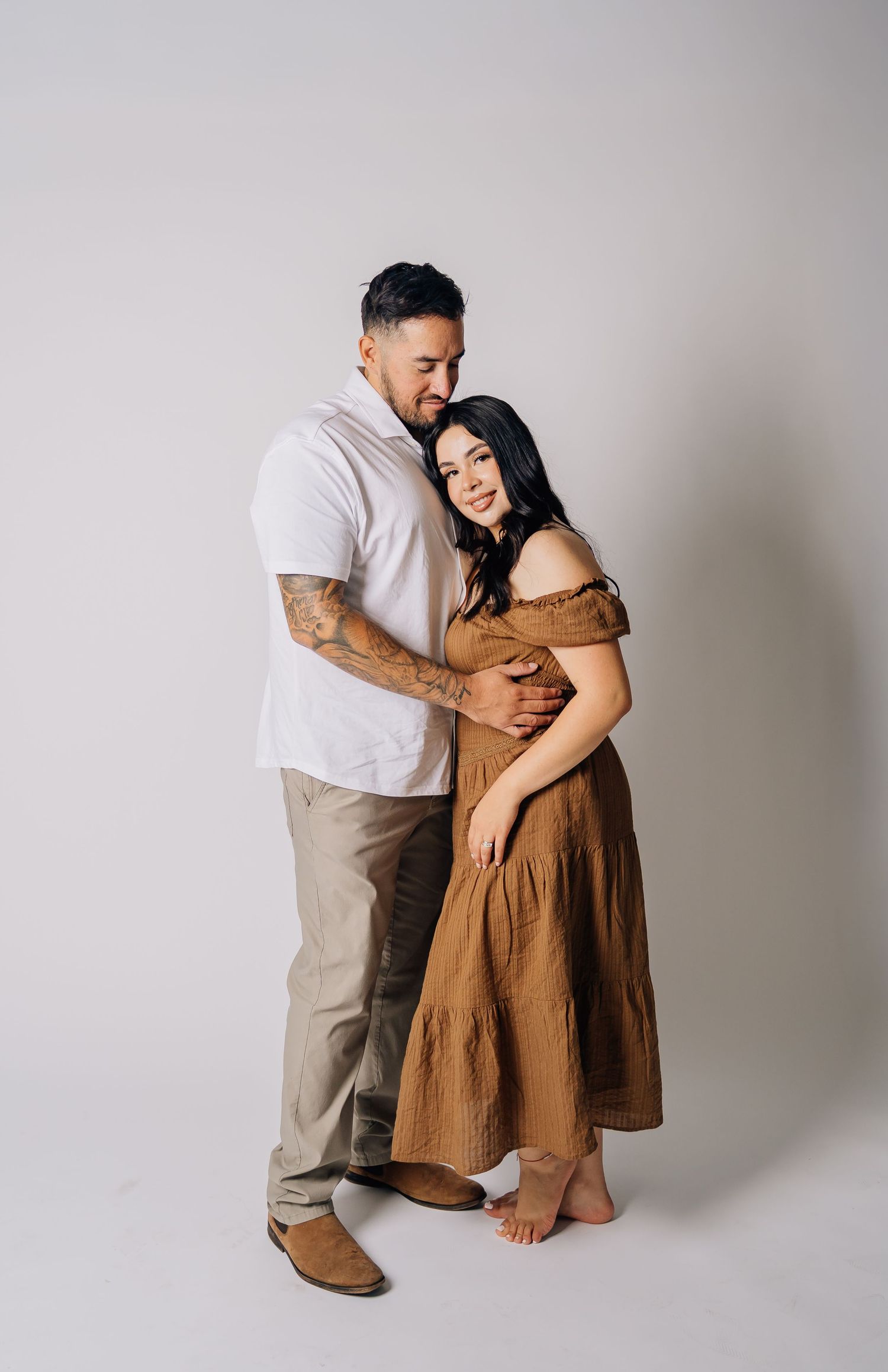 A couple embraces in a studio, man in white shirt with tattoos, woman in brown off-shoulder dress, against white background.
