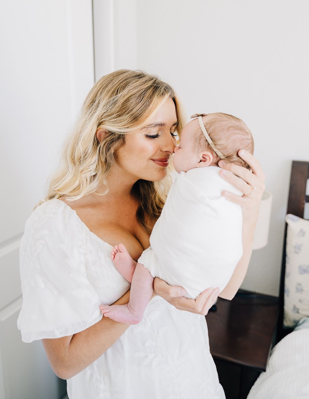 A blonde mother in a white dress tenderly kisses her newborn baby wrapped in white swaddle.