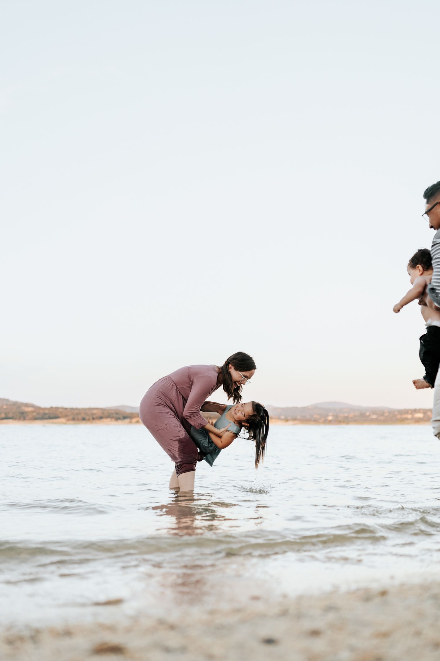 People playing and splashing in shallow water at the beach with hills in the background at sunset.