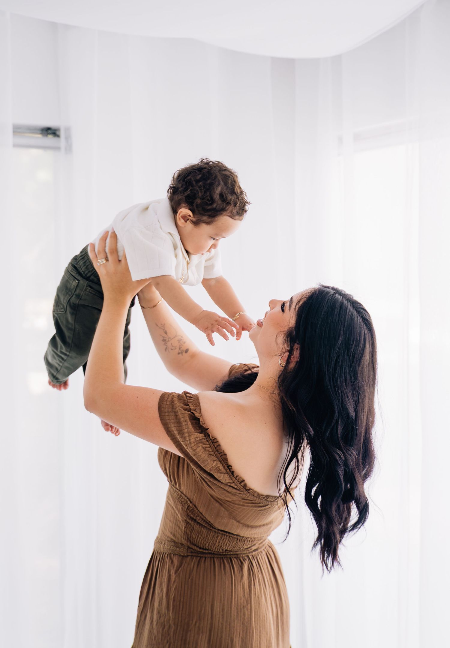 A happy mother in a brown dress lifts her baby overhead in an airy white studio setting.