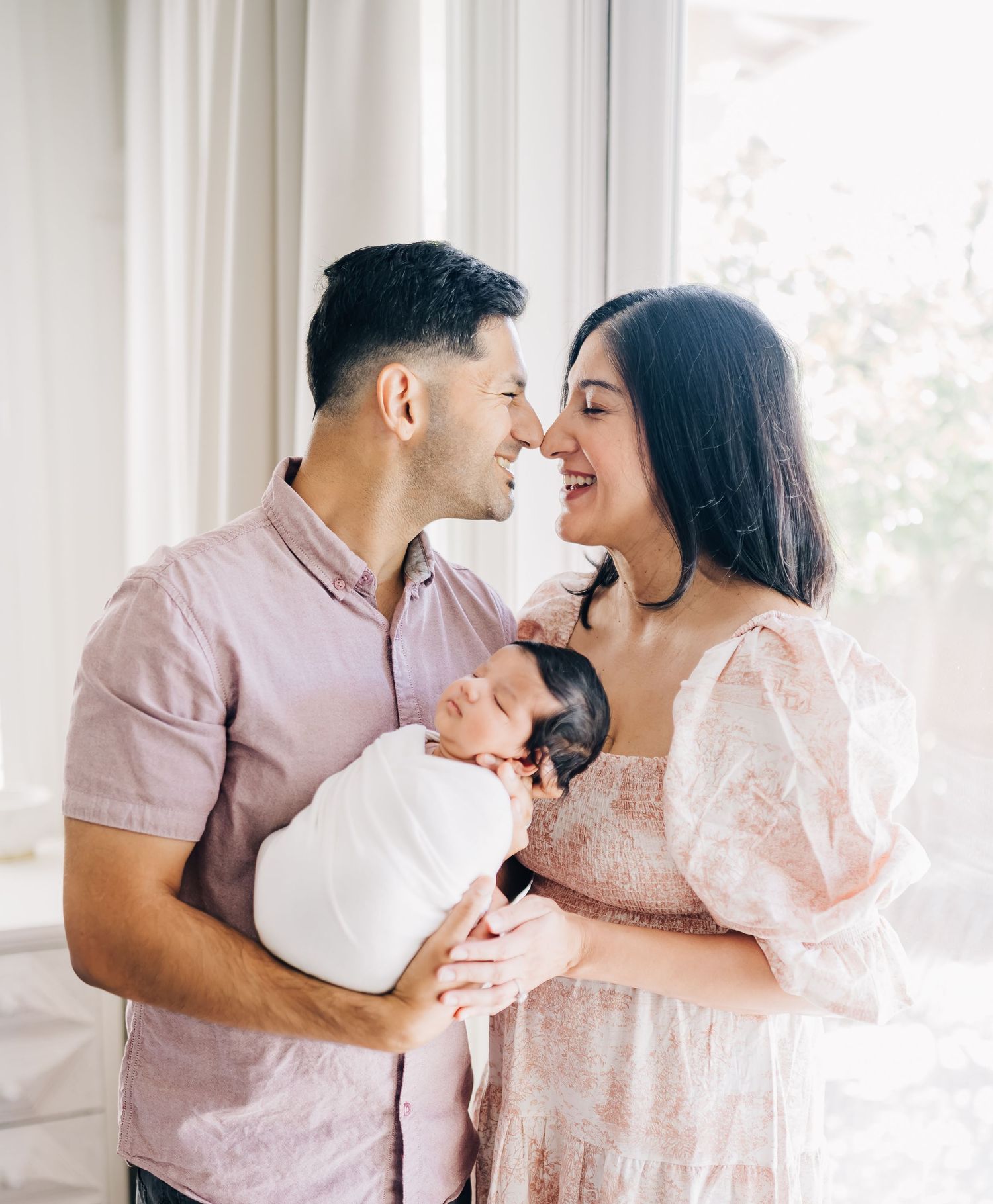 A happy couple smiles at each other while tenderly holding their swaddled newborn baby near a bright window.