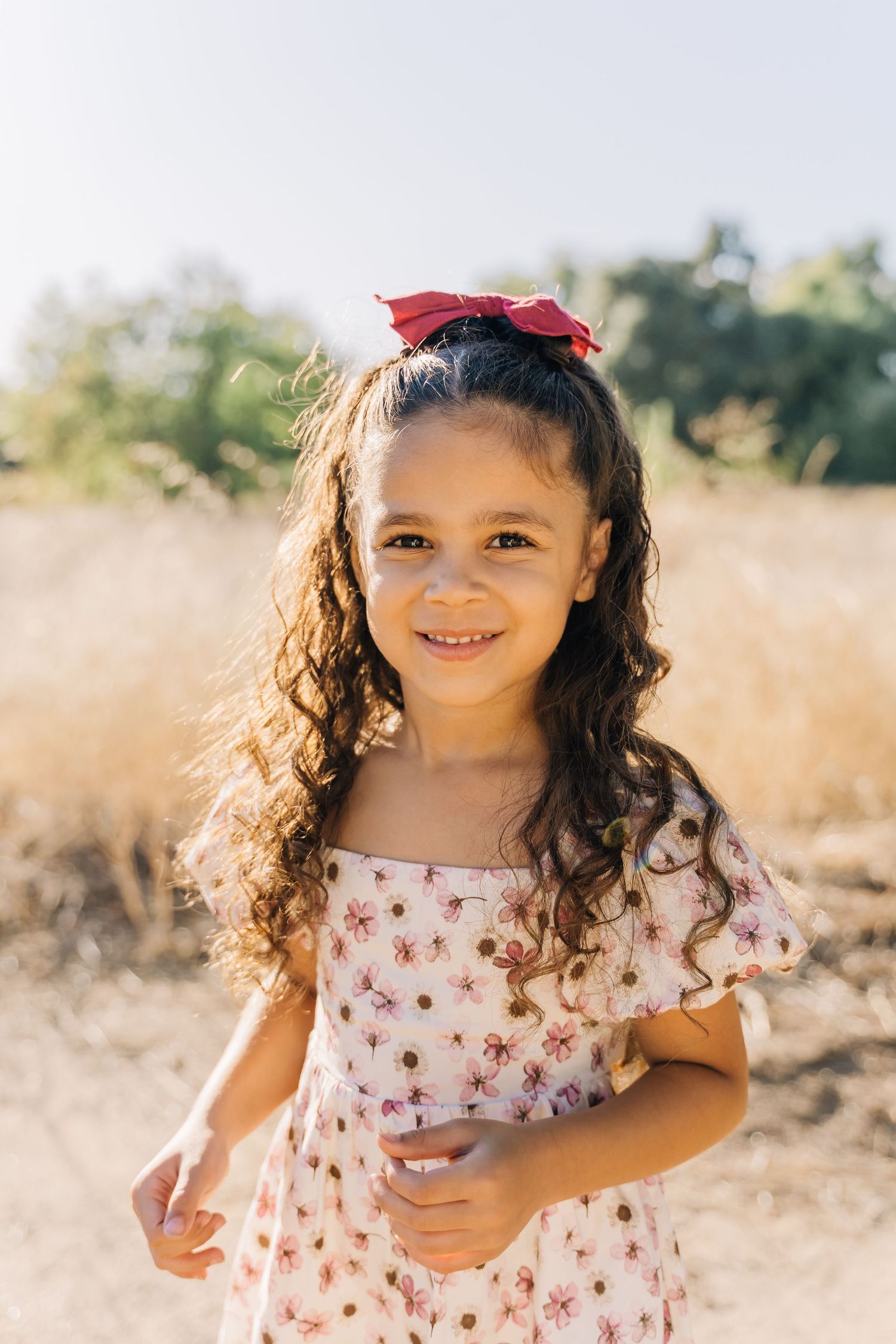 A young child in a floral dress plays and smiles in a sunny outdoor field setting.
