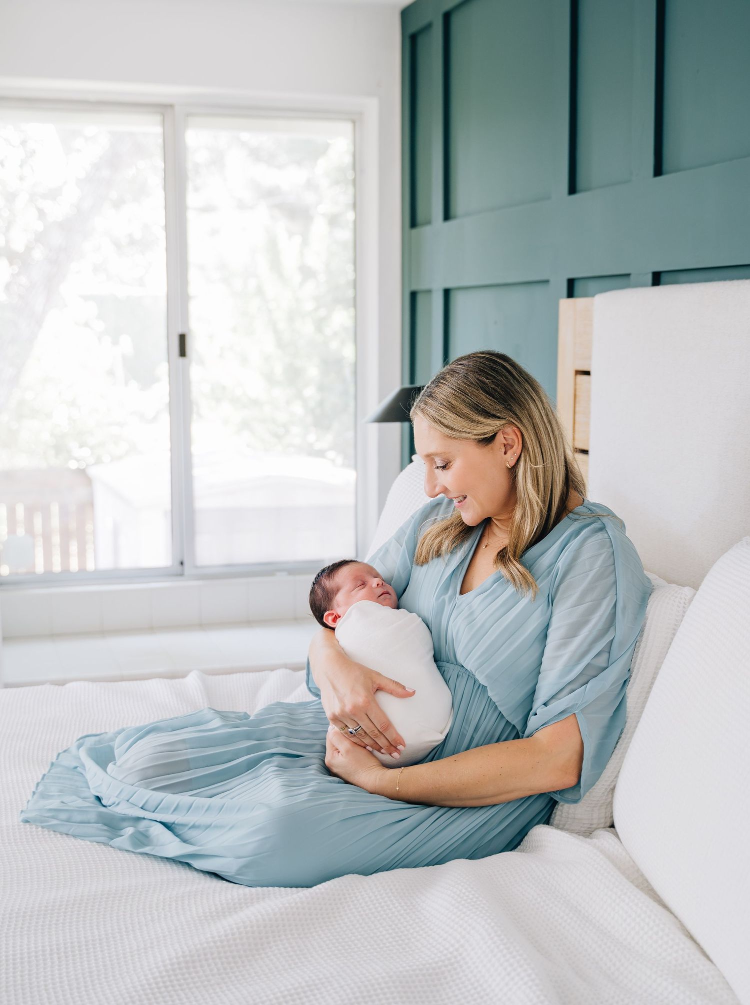A mother in a blue dress sits on a white bed, lovingly gazing at her swaddled newborn baby.