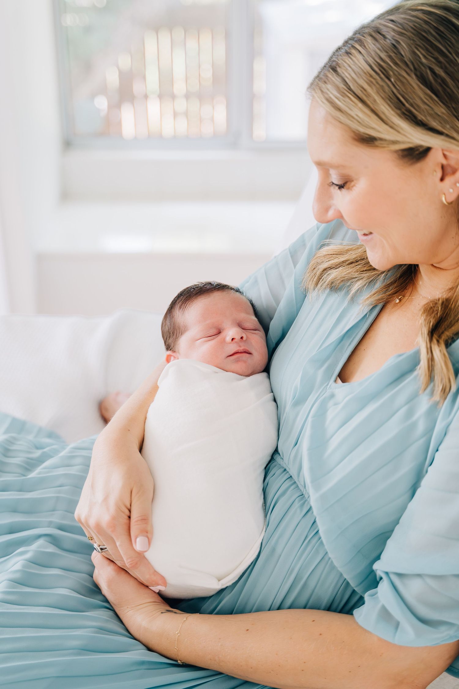 A mother in a light blue dress tenderly holds her sleeping swaddled newborn baby close to her chest.