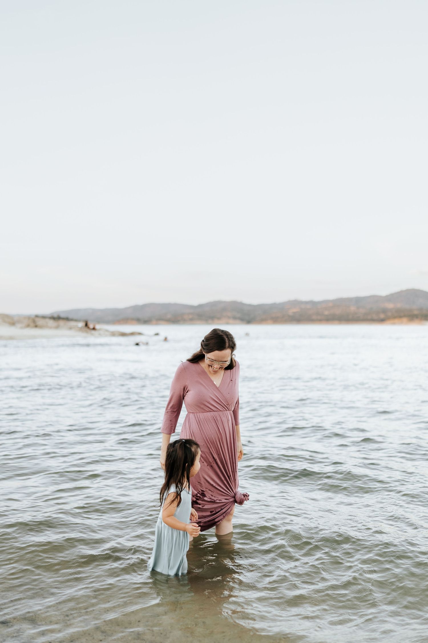 Two people wade together in shallow ocean water wearing casual clothing on a cloudy day at the beach.