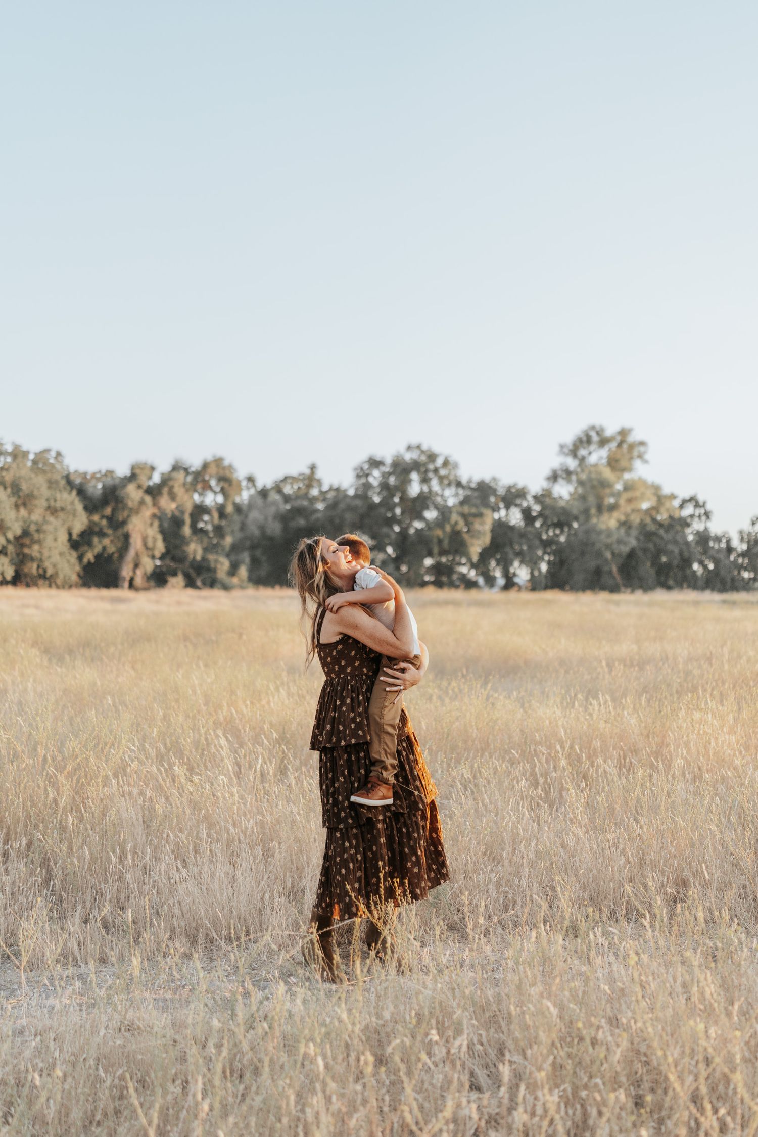 Two figures embrace tenderly in a golden field of tall grass at sunset with trees in the background.