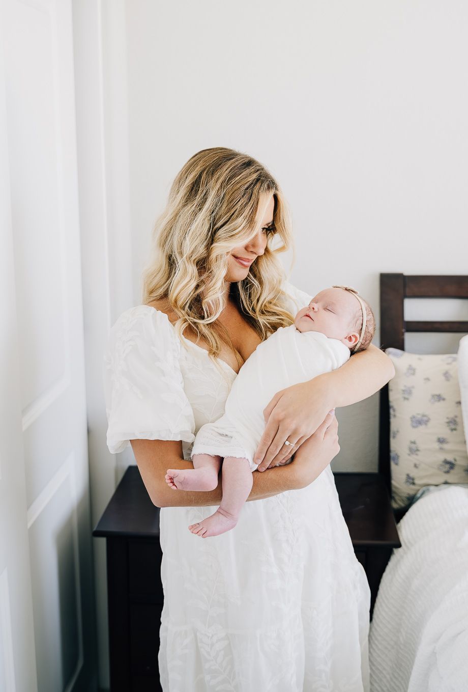 A mother in a white dress tenderly holds her sleeping newborn baby while standing in a softly lit bedroom.