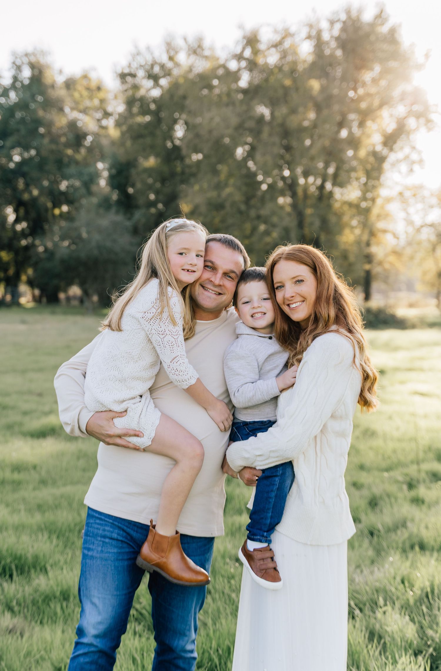 A family in coordinating white tops and jeans embraces while posing in a sunlit grassy field during golden hour.