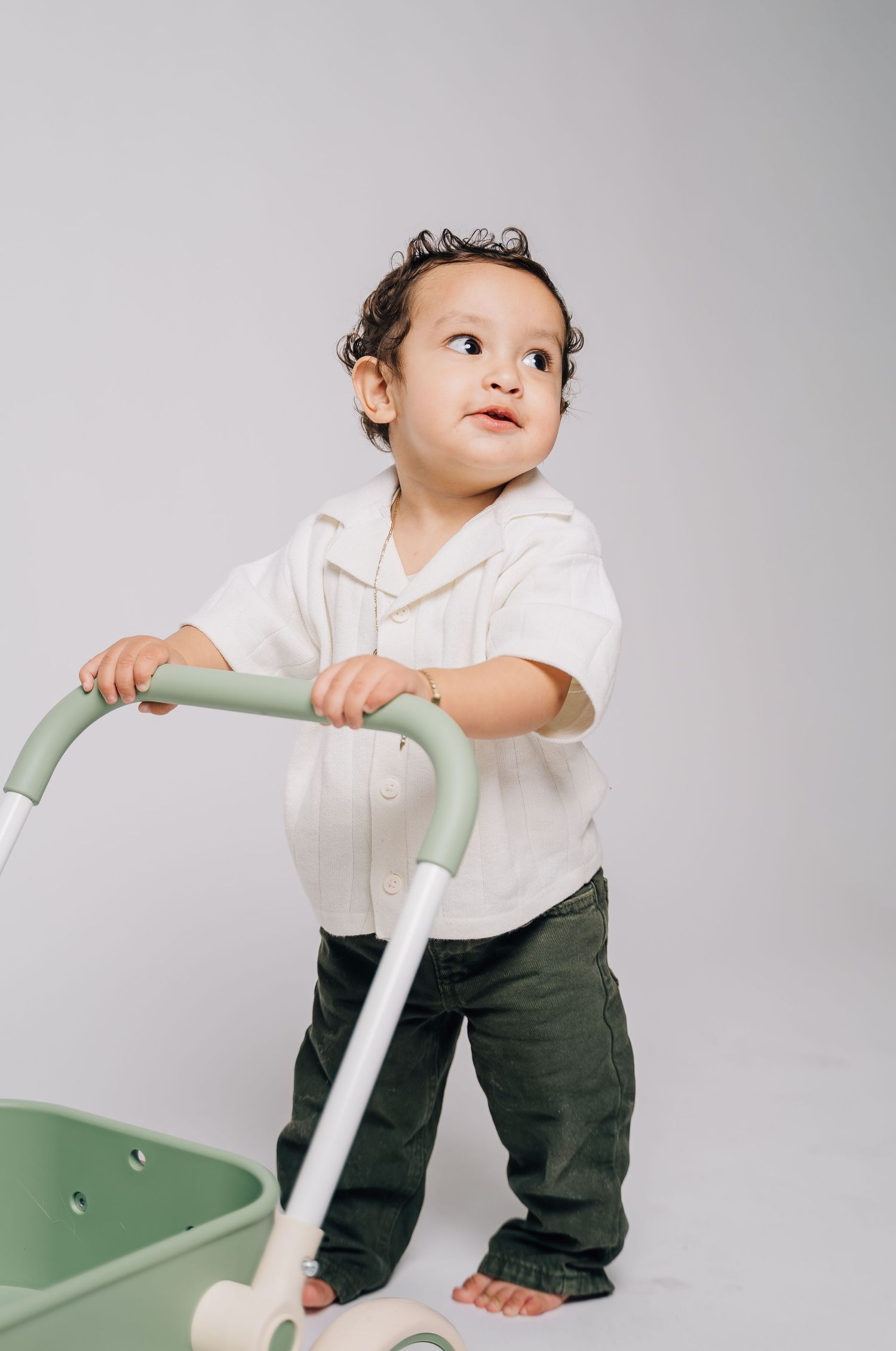 Toddler in white shirt and green pants pushing a mint green baby walker against a white background.