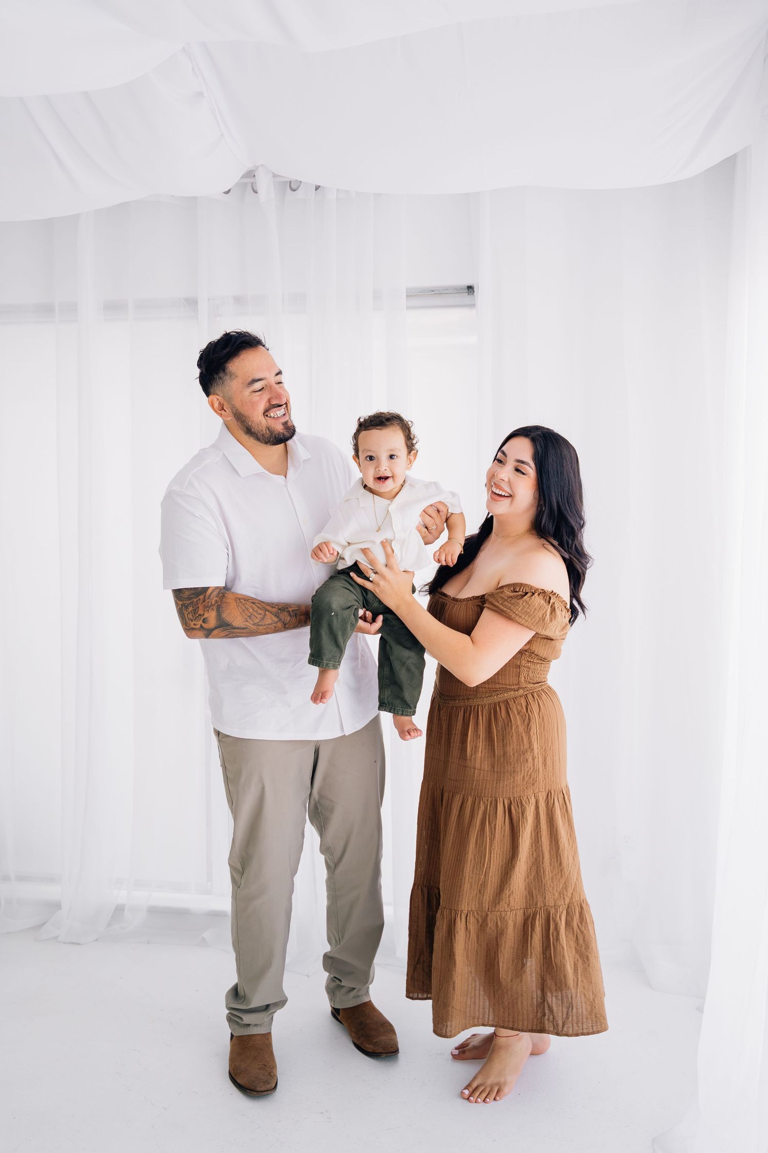 Happy family portrait with tattooed father, mother in brown dress, and smiling baby in bright white studio.