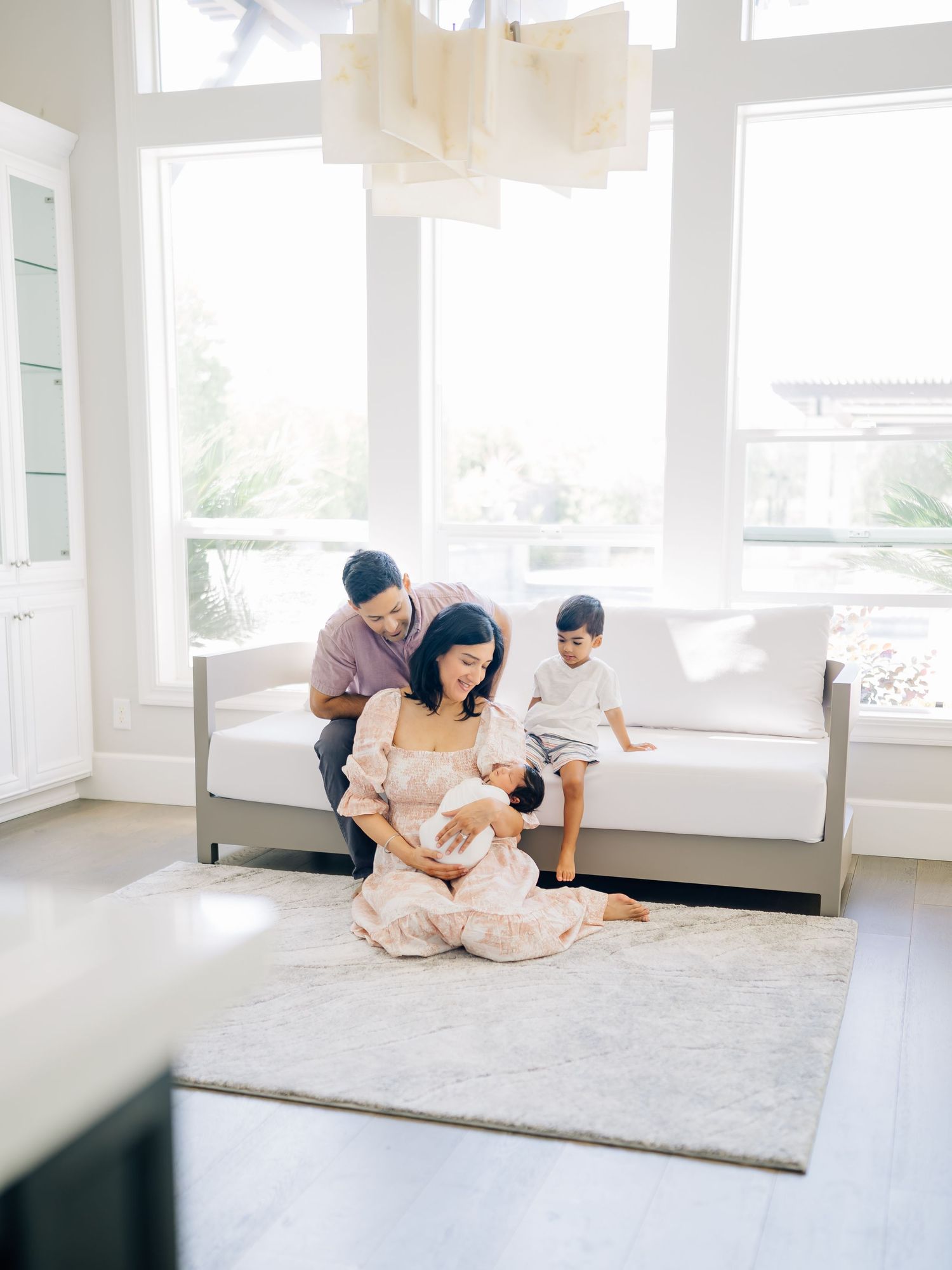 A family of four sits together in a bright, airy living room, with a mother cradling a newborn baby.