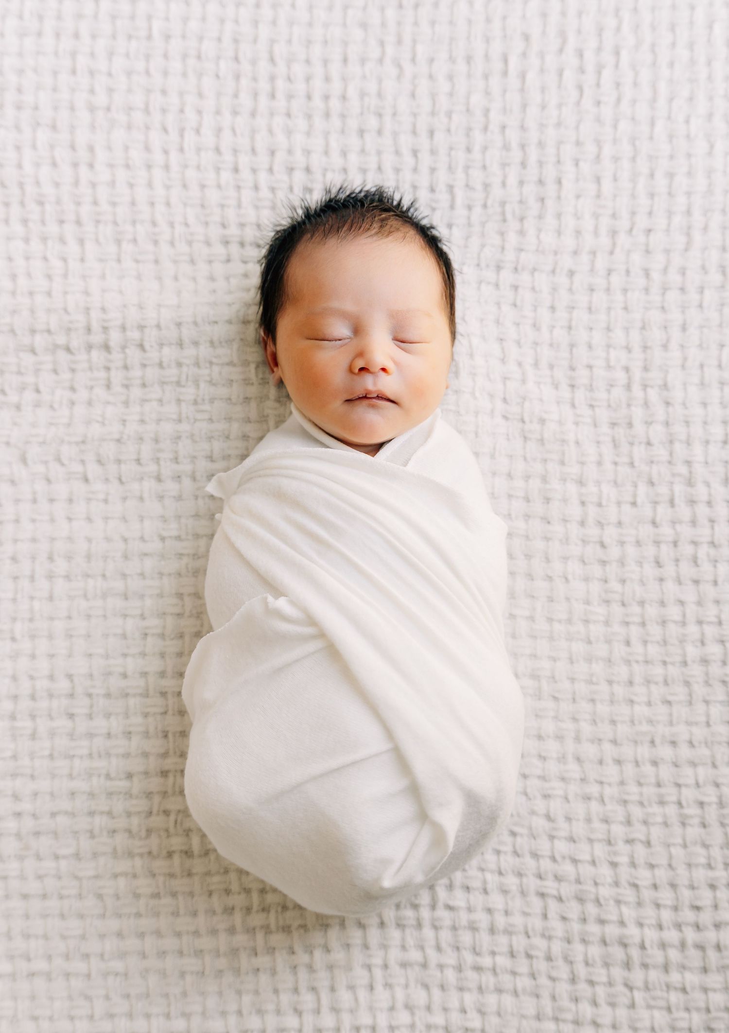 Peaceful newborn baby swaddled in a white wrap, sleeping on a soft white textured blanket.
