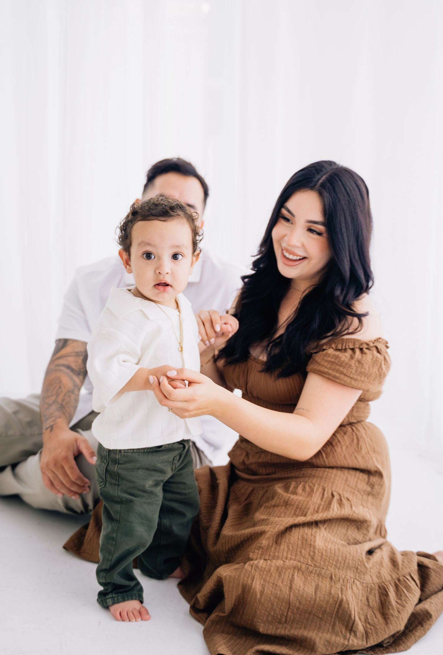A family with a toddler boy poses in a bright white studio, mom in brown dress smiling warmly.