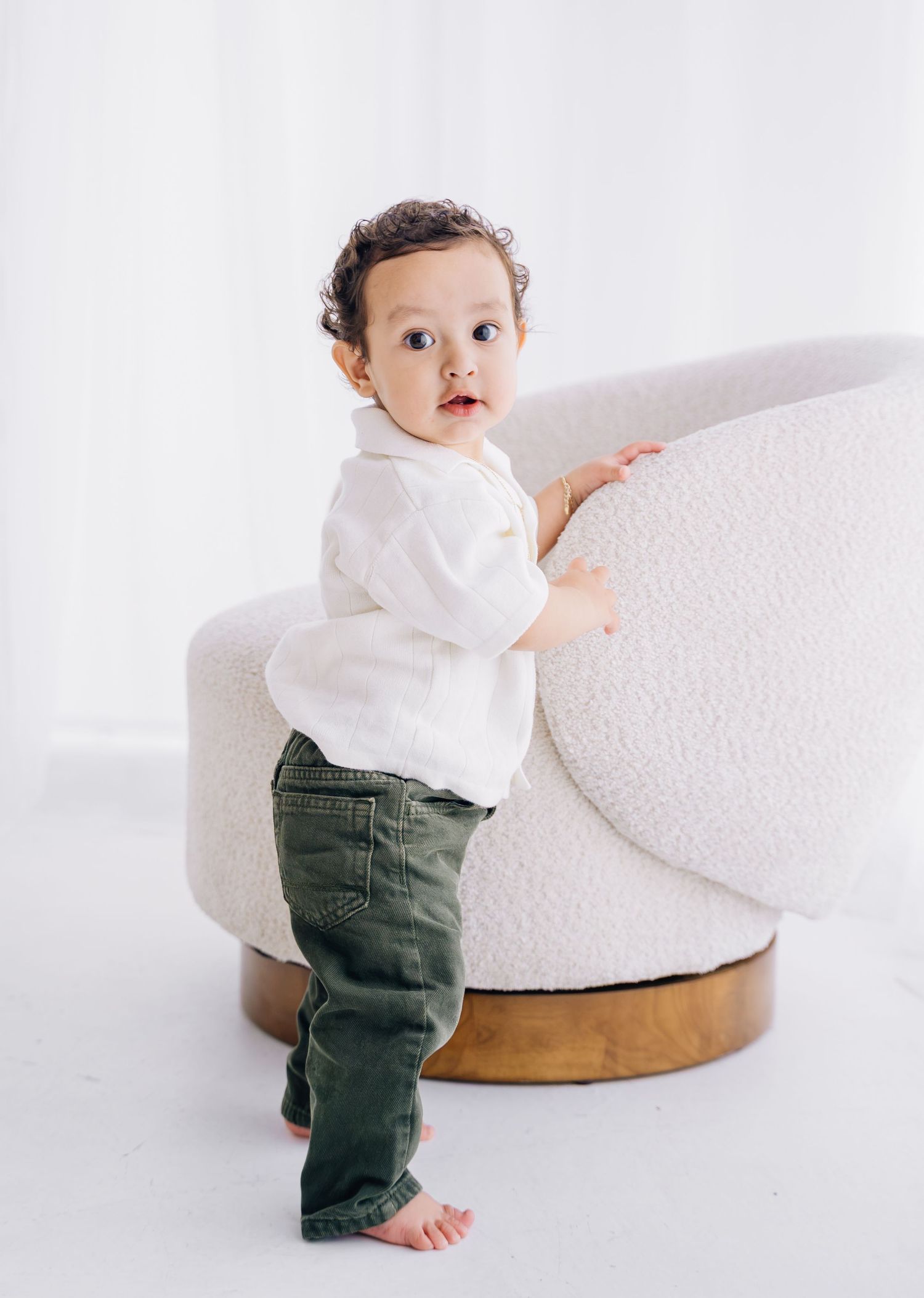 Adorable toddler in white shirt and green pants standing by a cream boucle chair in a bright white studio.