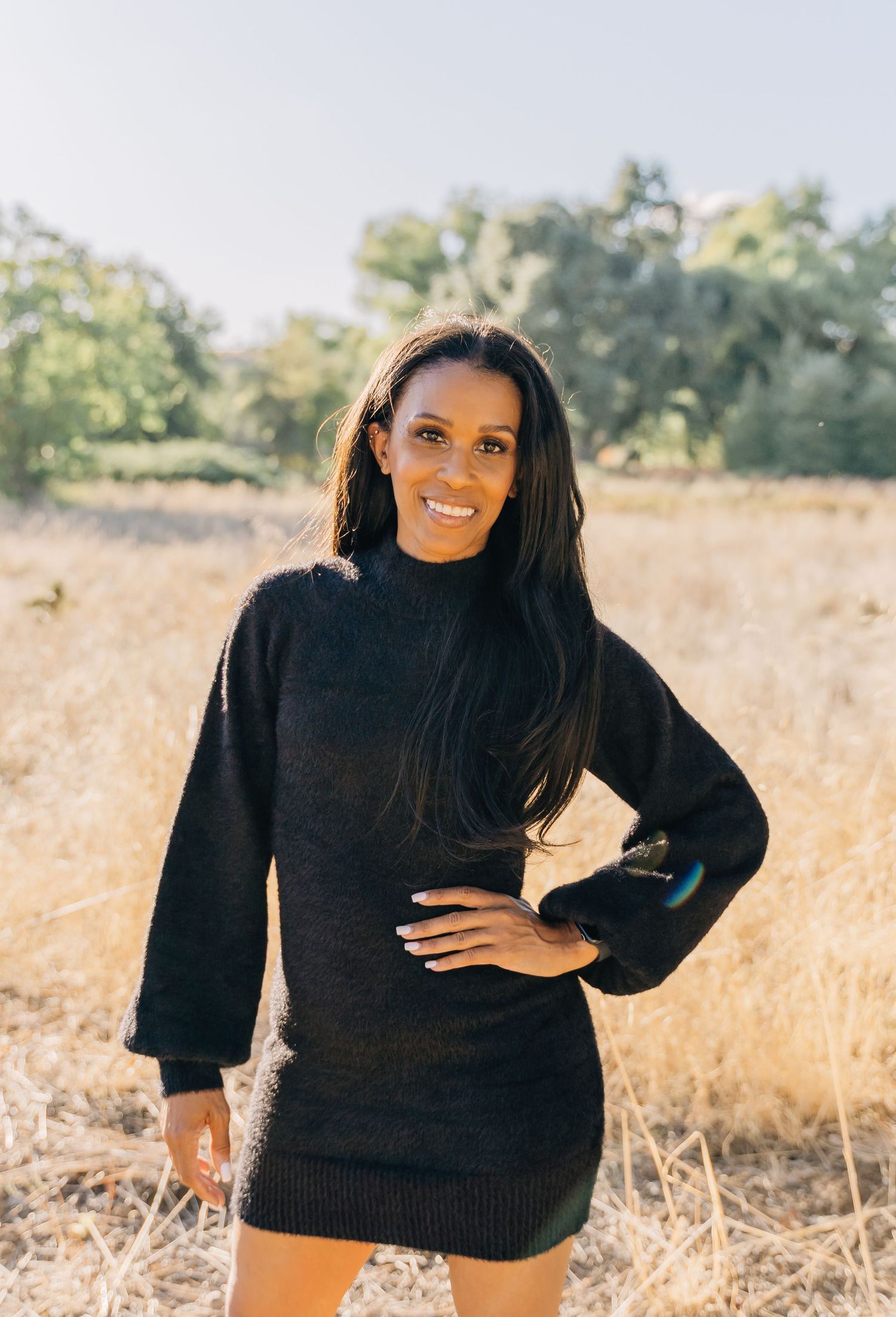 Someone wearing a cozy black sweater dress poses outdoors in a golden field on a sunny autumn day.