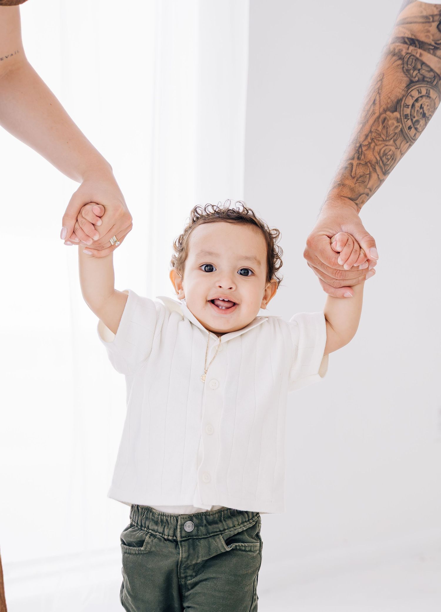 A happy toddler in white shirt and green pants stands holding both parents hands against white background.