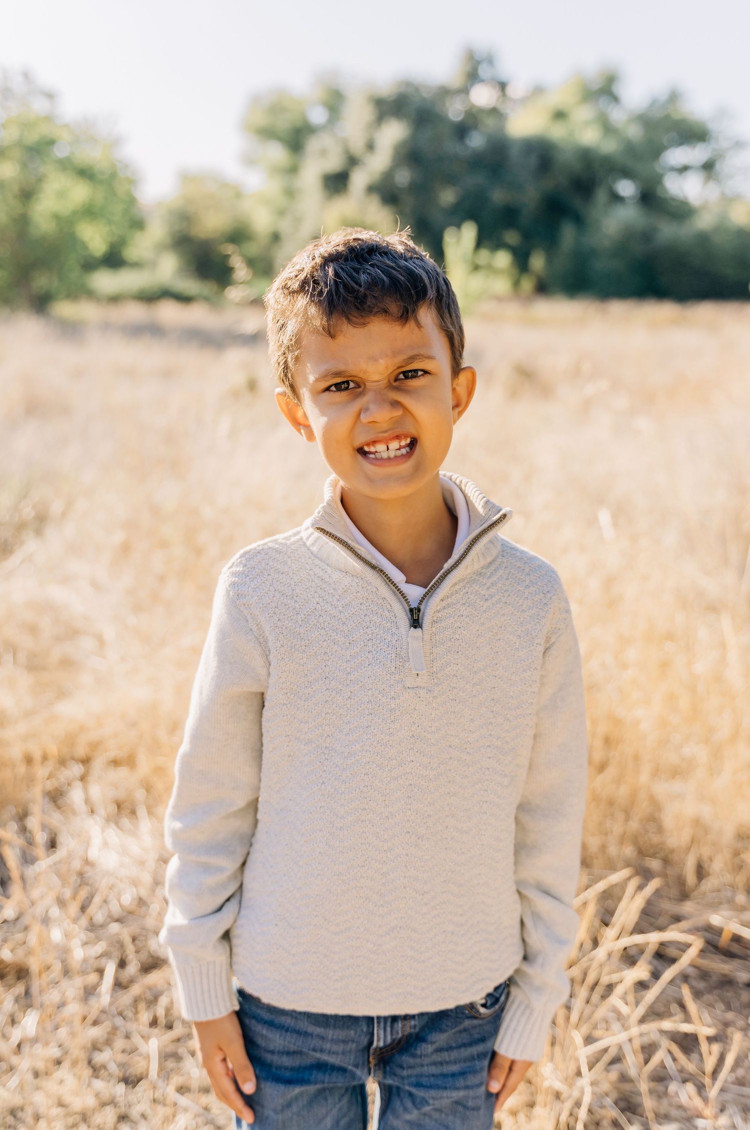 A child wearing a cream colored sweater poses outdoors in a sunny field with dried grass.