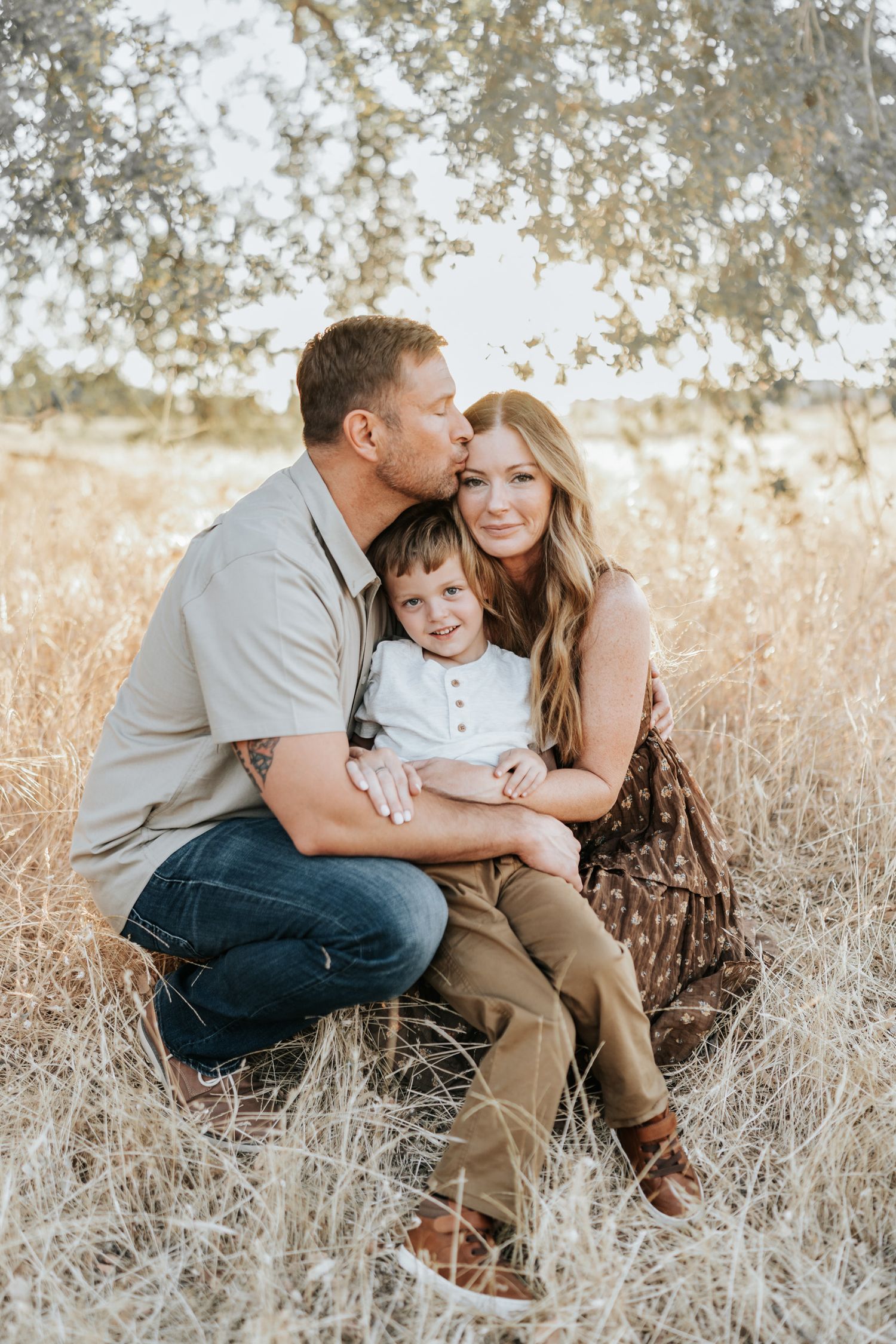 Family of three sharing tender moments together in golden sunlit field of tall grass and wildflowers.