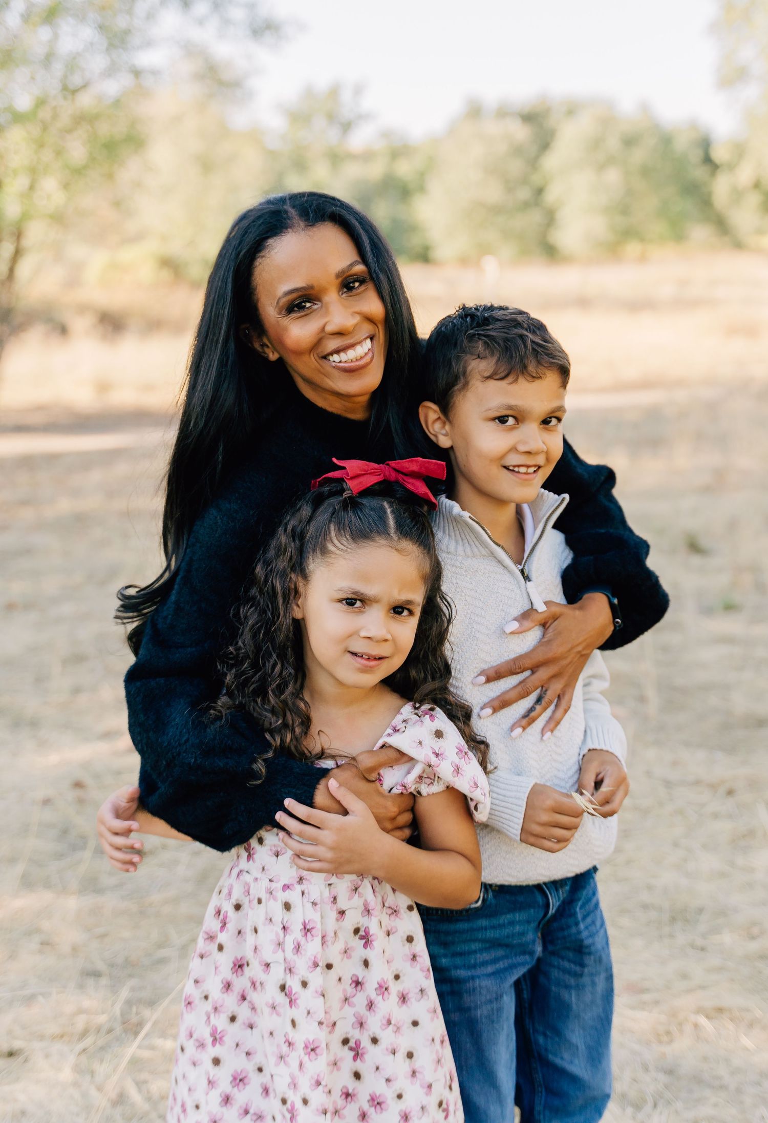 A loving family moment captured outdoors with a mother embracing her two children on a sunny day.