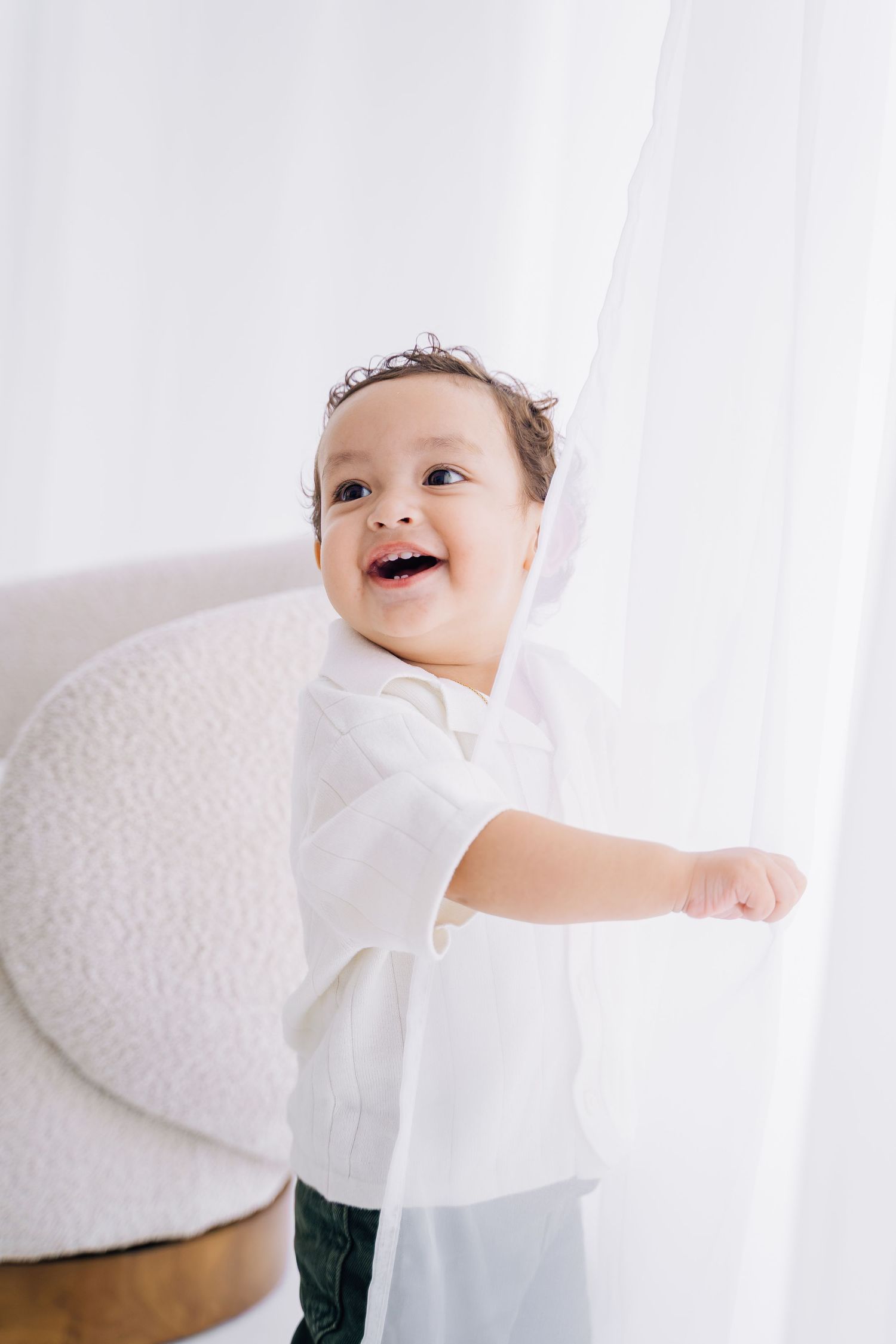 Happy baby in white outfit laughing while holding a sheer curtain in a bright, airy room.