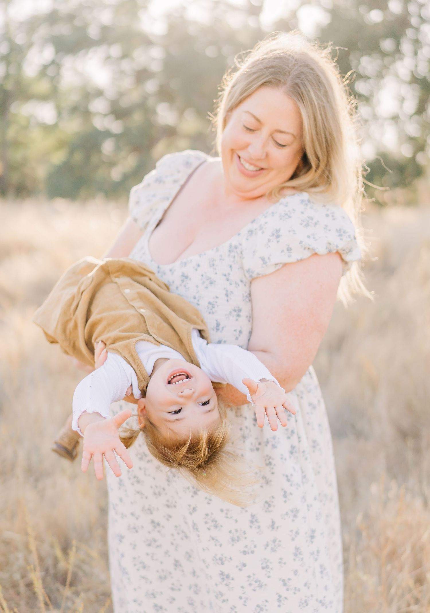 A smiling mother in a floral dress playfully holds her laughing toddler upside down in a sunlit field.