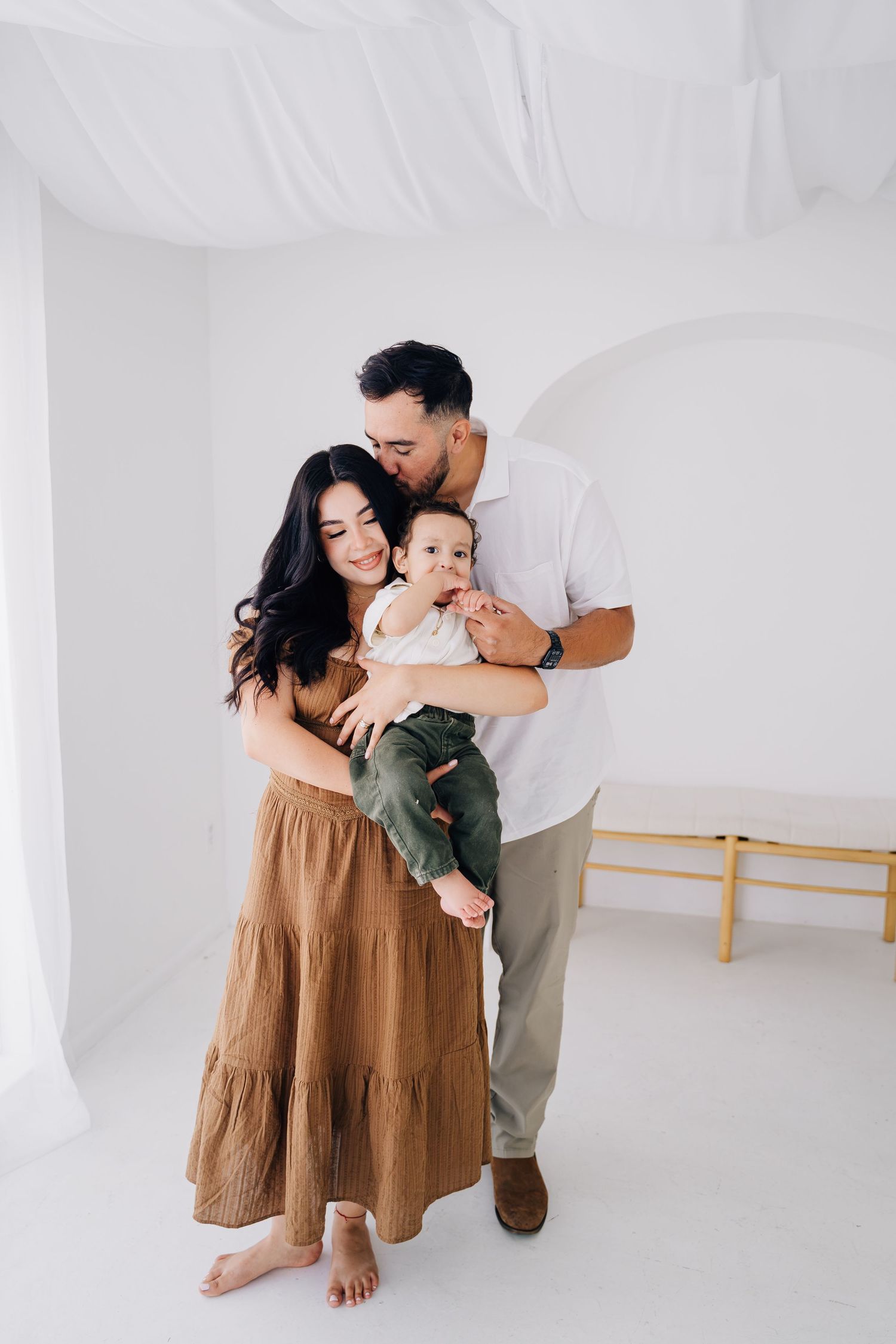 A young family poses in a white studio, mother in tan dress holds baby while father kisses her head.