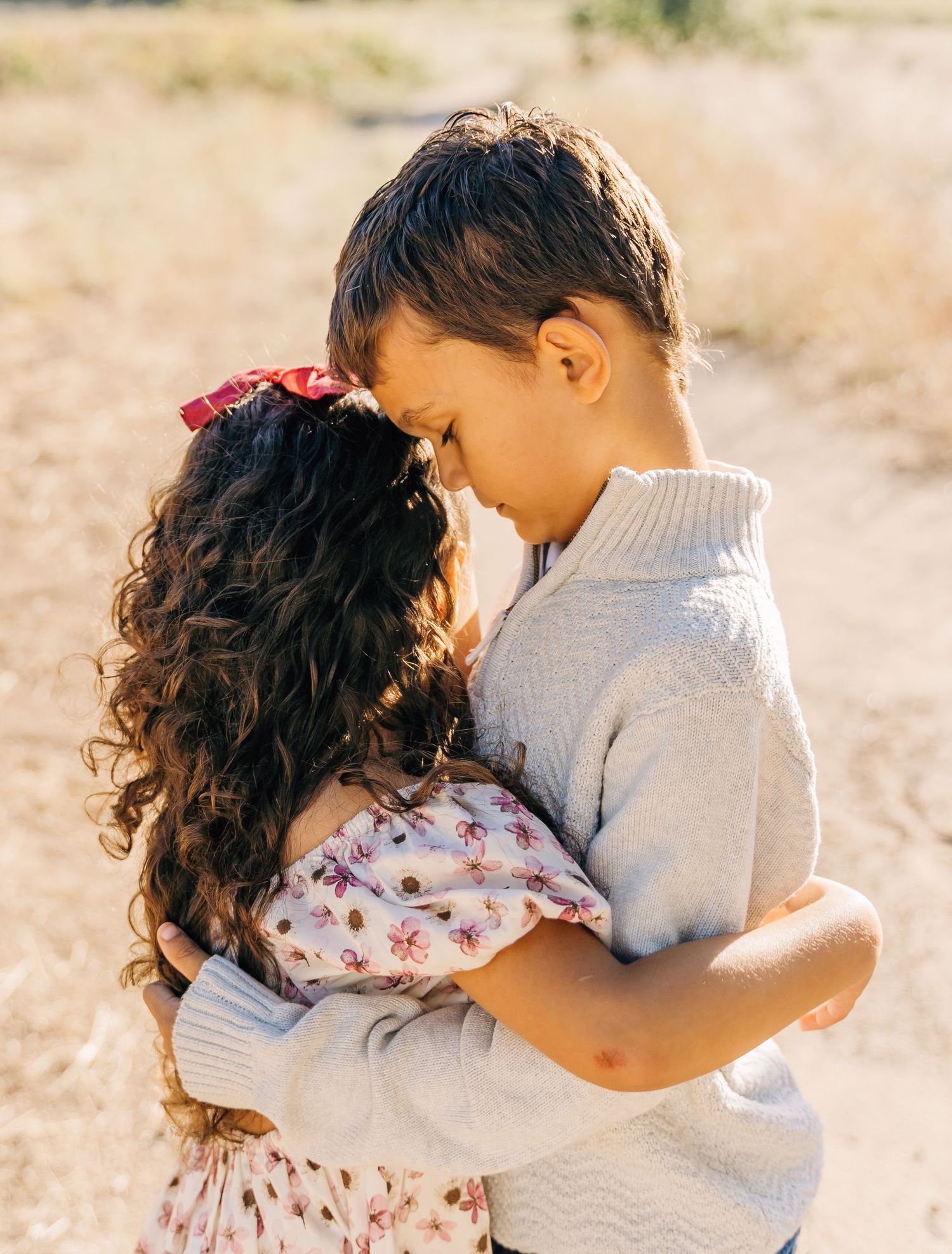 Two young children share a tender embrace while standing together in a sunlit outdoor setting.
