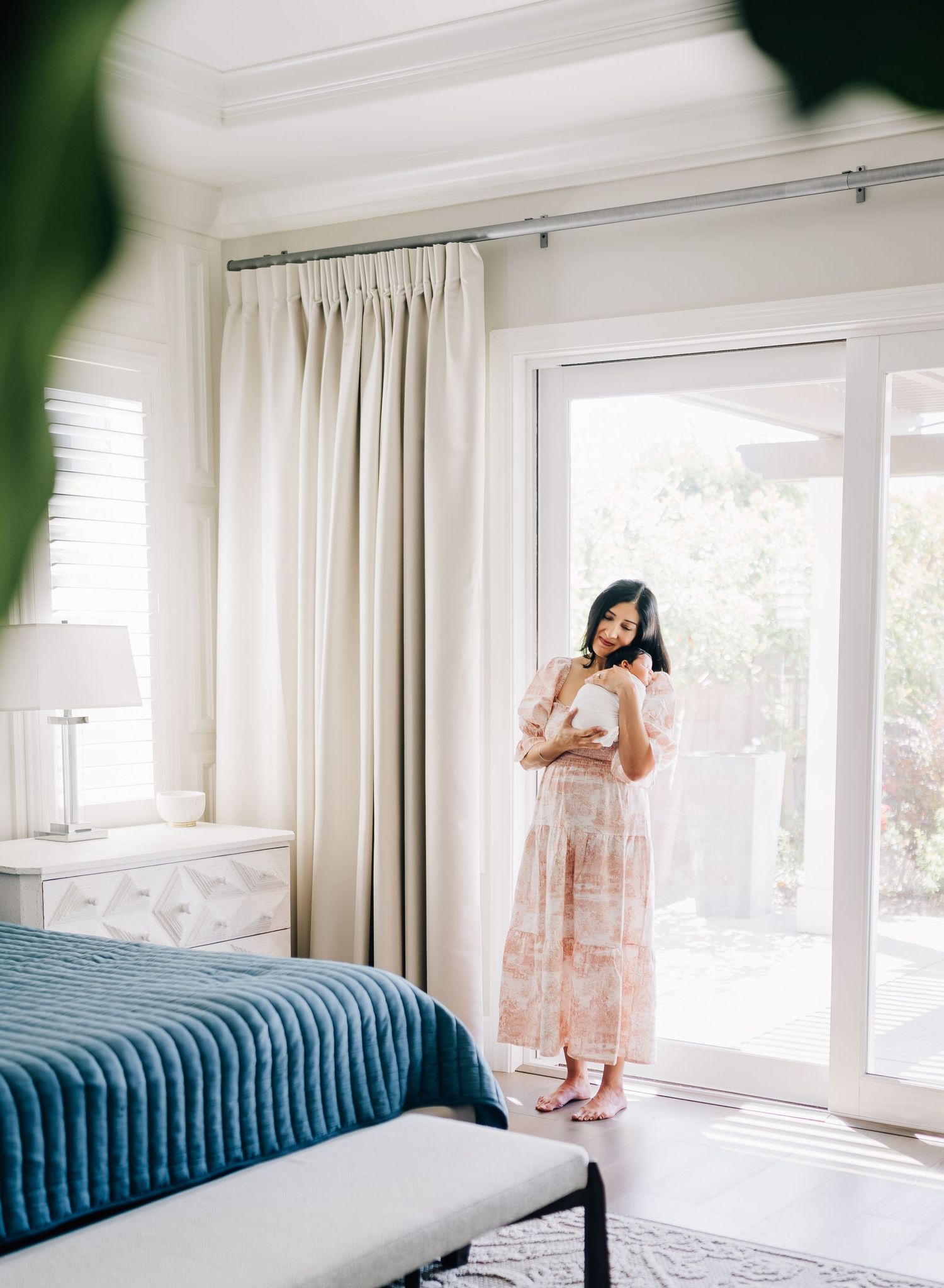 Woman in pink floral maxi dress holding a bowl near sliding glass doors in a bright, elegant bedroom.