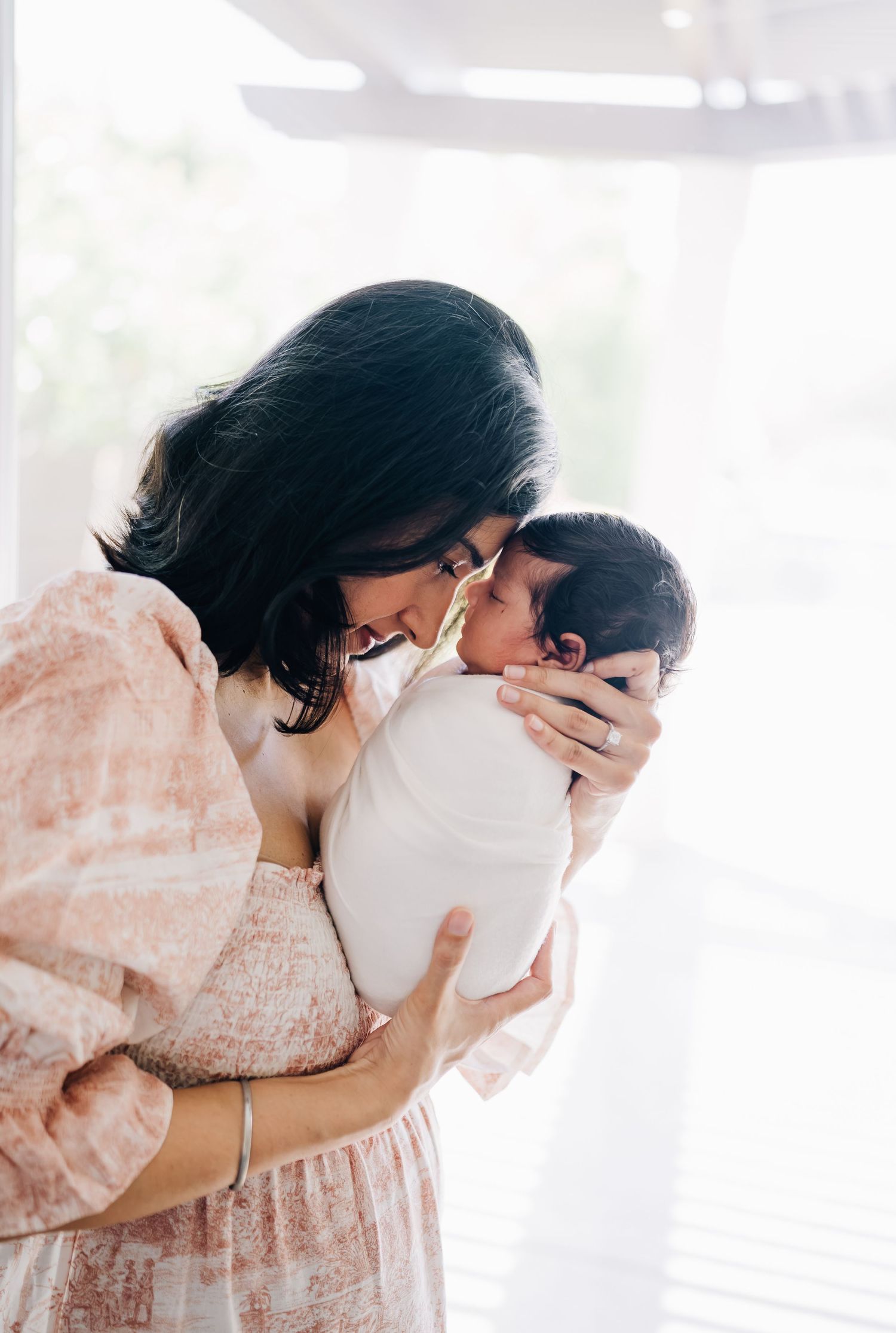 A mother tenderly nuzzles her newborn baby wrapped in white, sharing a precious bonding moment near a bright window.