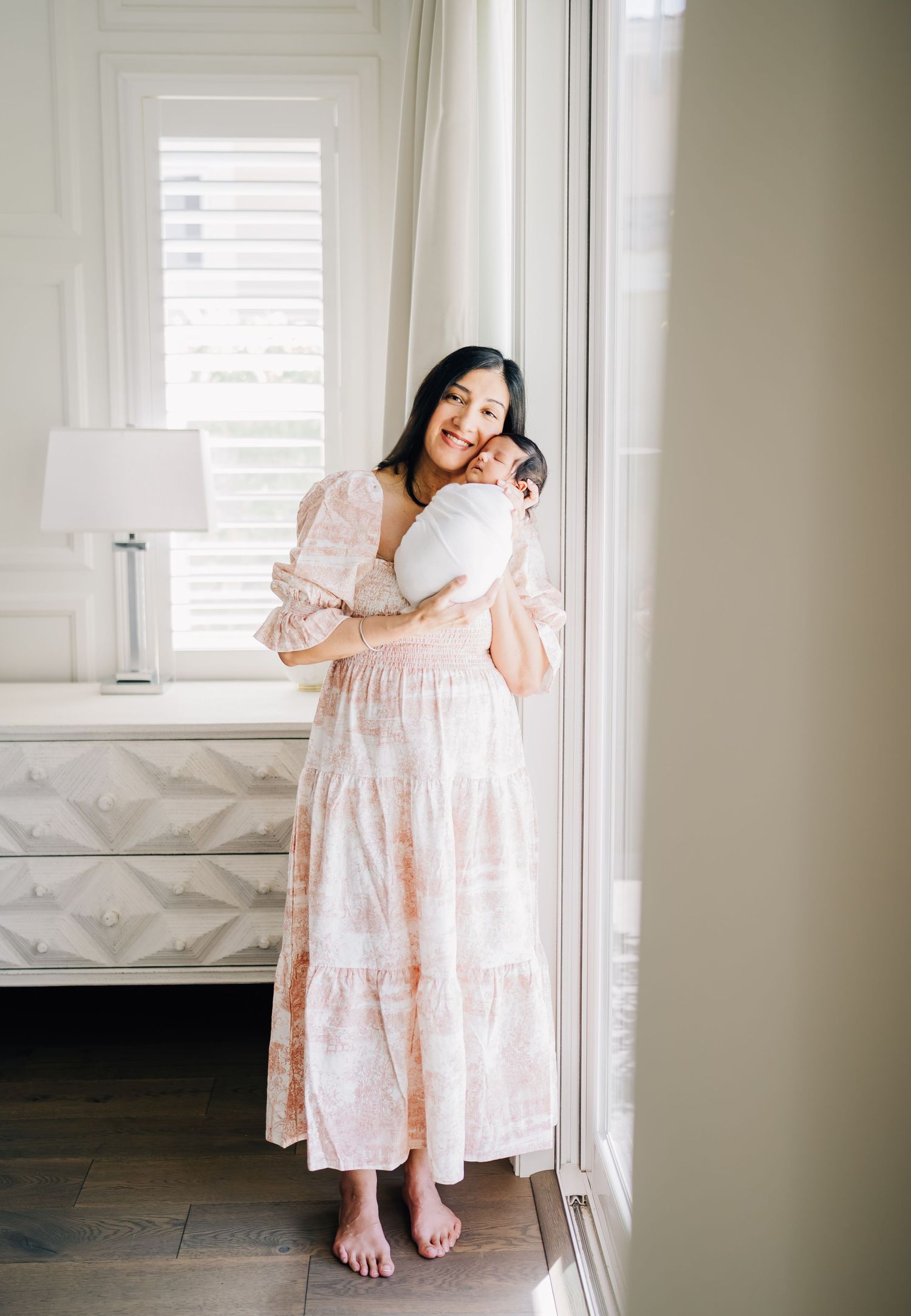 A woman in a pink floral dress holds a newborn baby near a bright window in an elegant white bedroom.