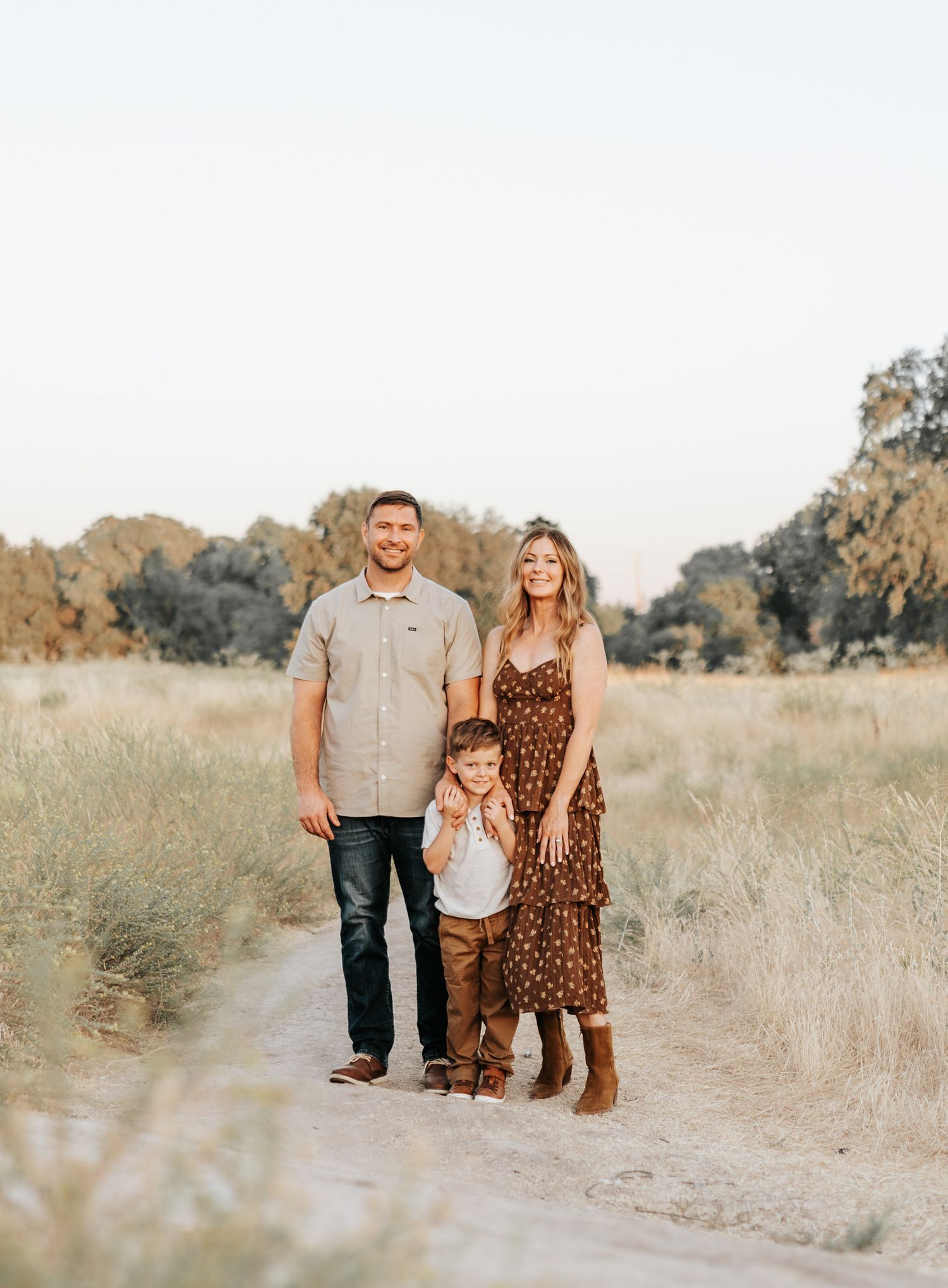 A family poses together on a dirt path surrounded by dry grass and trees during golden hour.