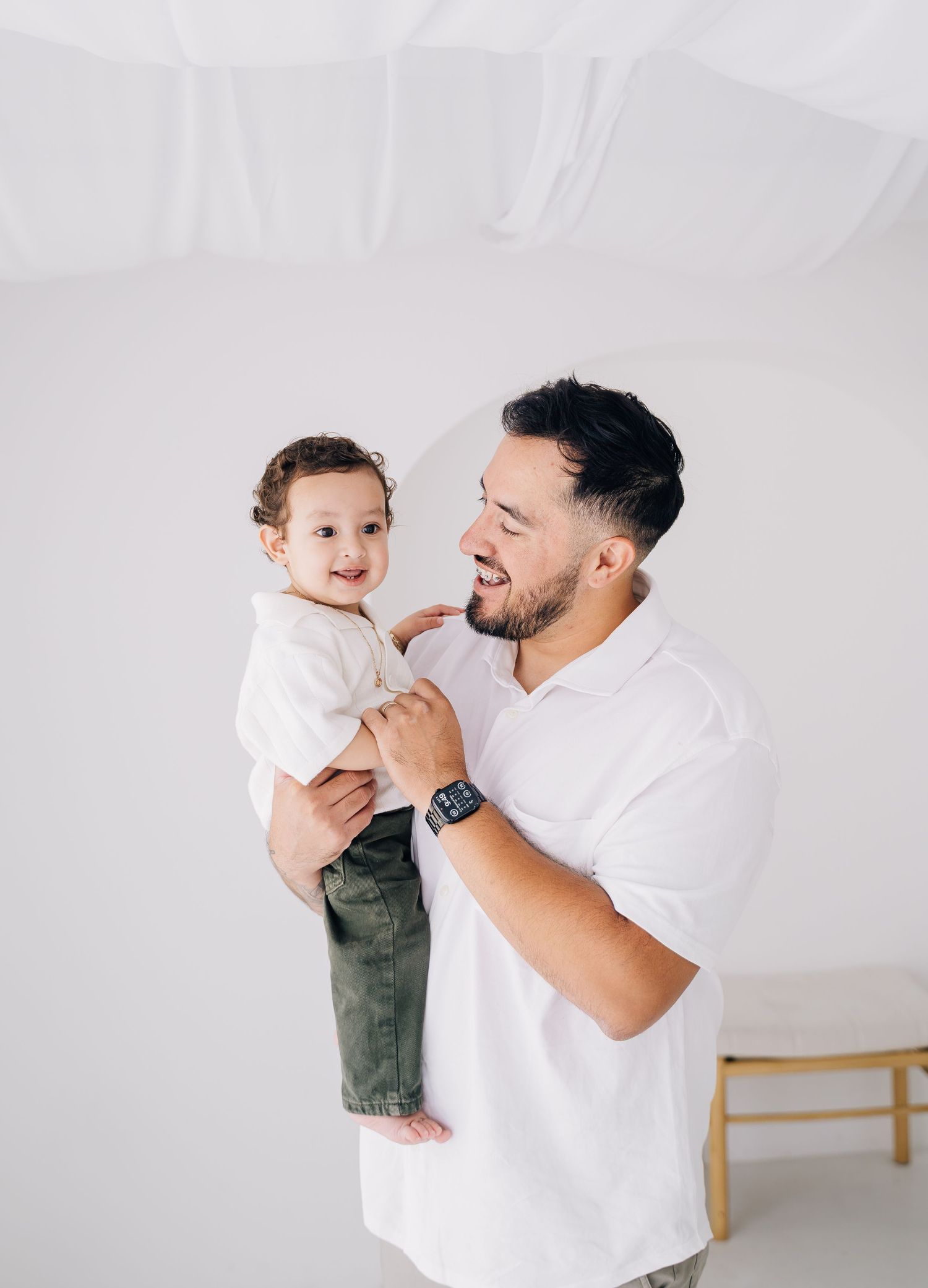 A smiling man in a white polo holds a happy toddler in olive pants against a bright white background.