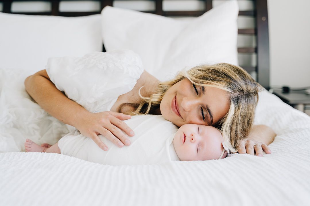 A smiling blonde mother lying on a white bed gazing lovingly at her sleeping newborn baby wrapped in white.