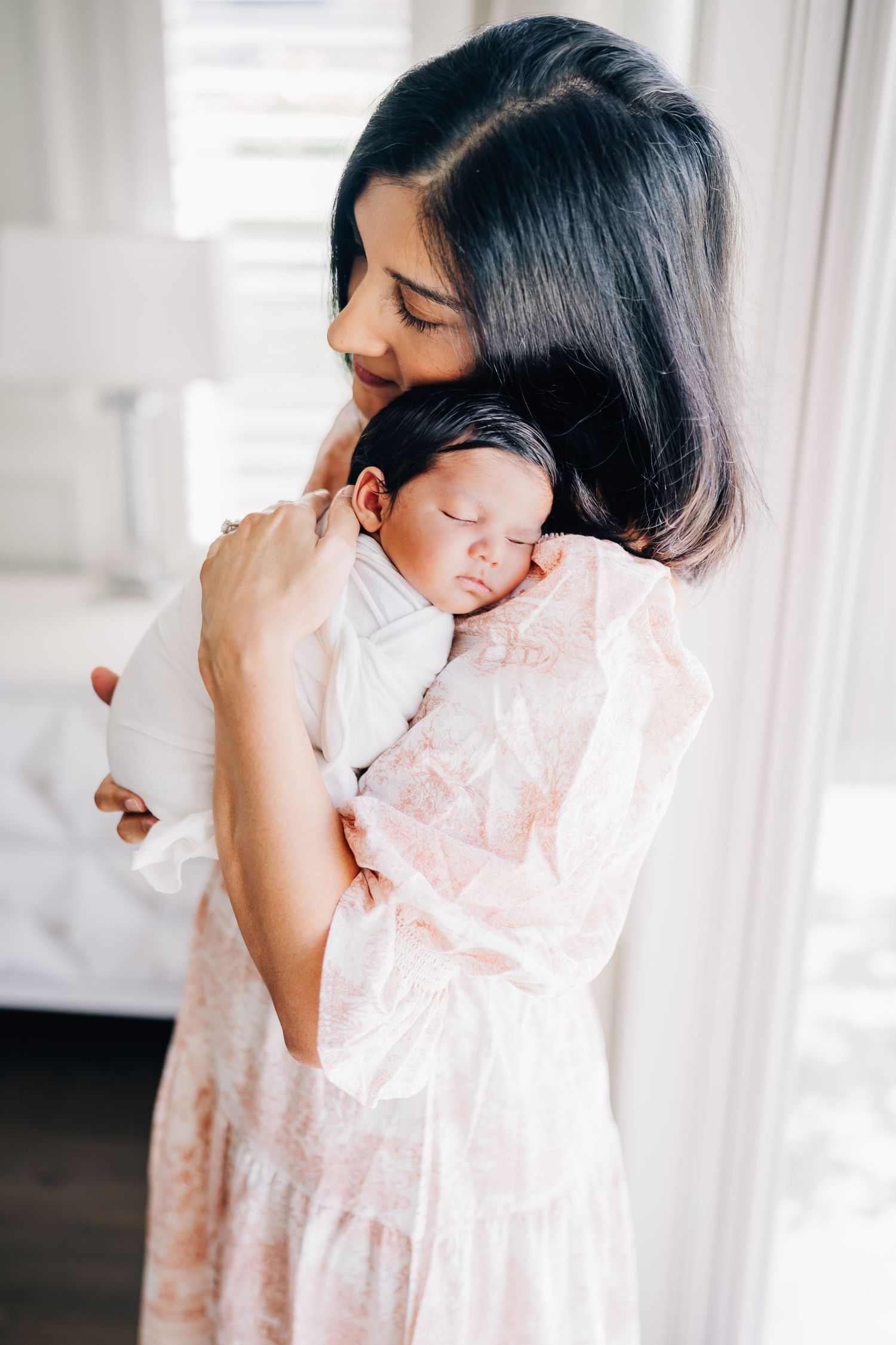 A mother in a floral dress tenderly holds her sleeping newborn baby against her chest in a bright, airy room.
