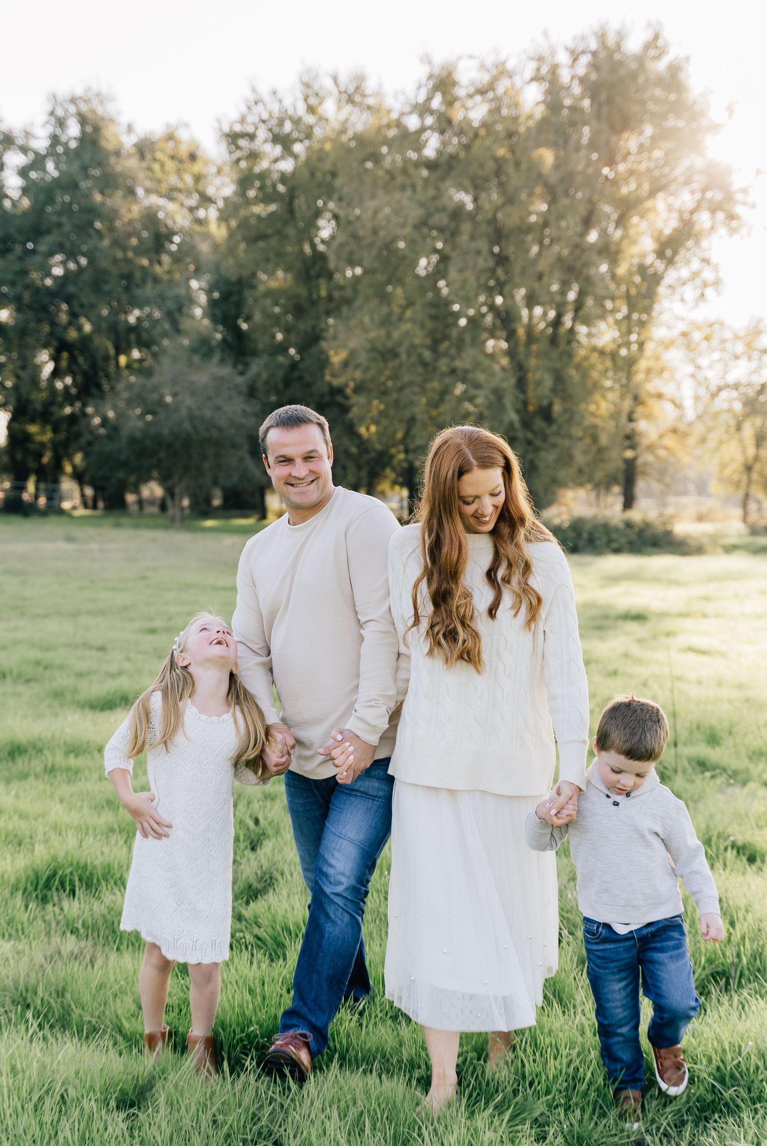 Family in coordinating white and denim outfits walking together in a sunny green field at sunset.