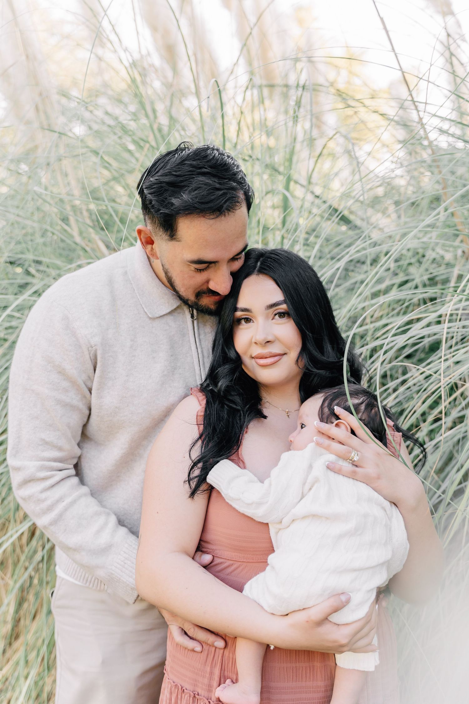 A couple tenderly holds their newborn baby outdoors among tall grasses, the man kissing the woman's head.