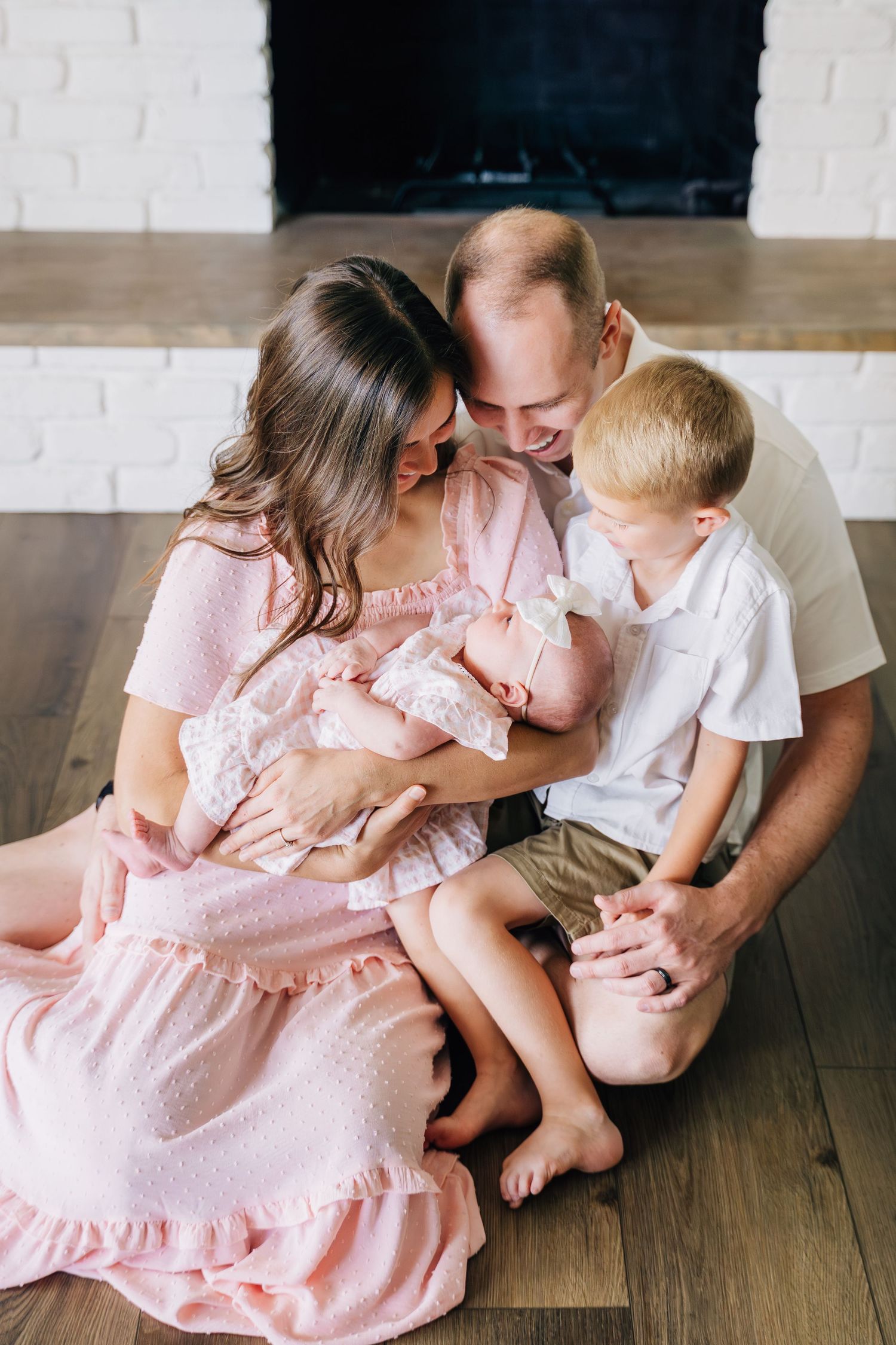 A loving family of four sits on hardwood floor, parents admiring newborn baby girl in pink while toddler son looks on.
