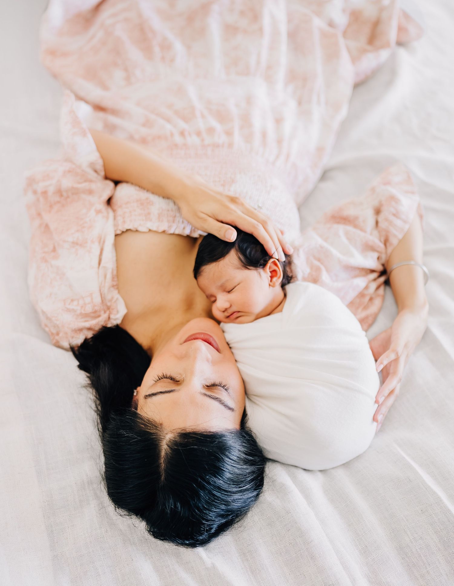 A mother in a pink floral dress lies on white bedding, cradling her sleeping newborn baby against her cheek.