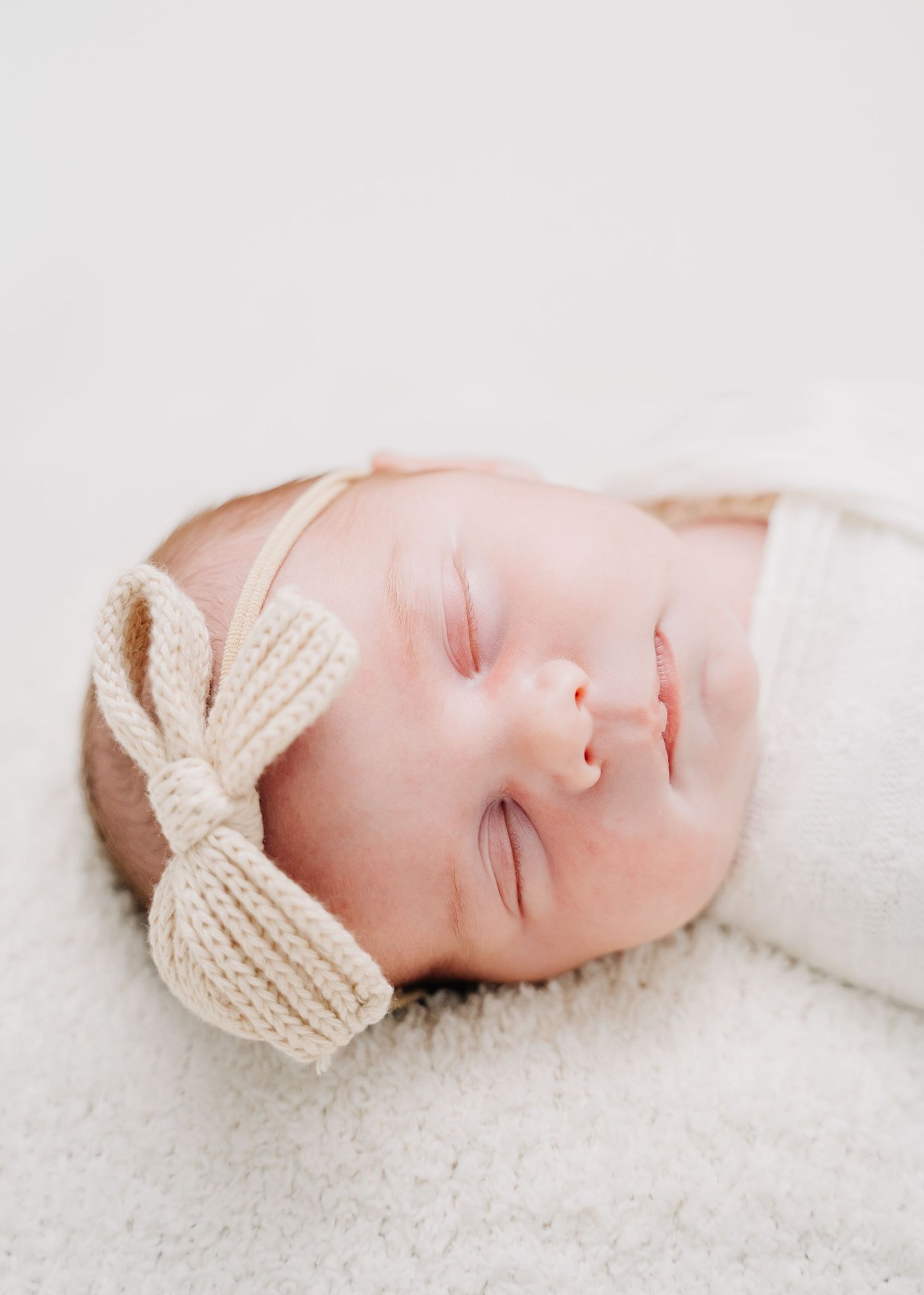 Sleeping newborn baby wearing a cream knit bow headband, swaddled in white, lying on a soft fluffy white blanket.