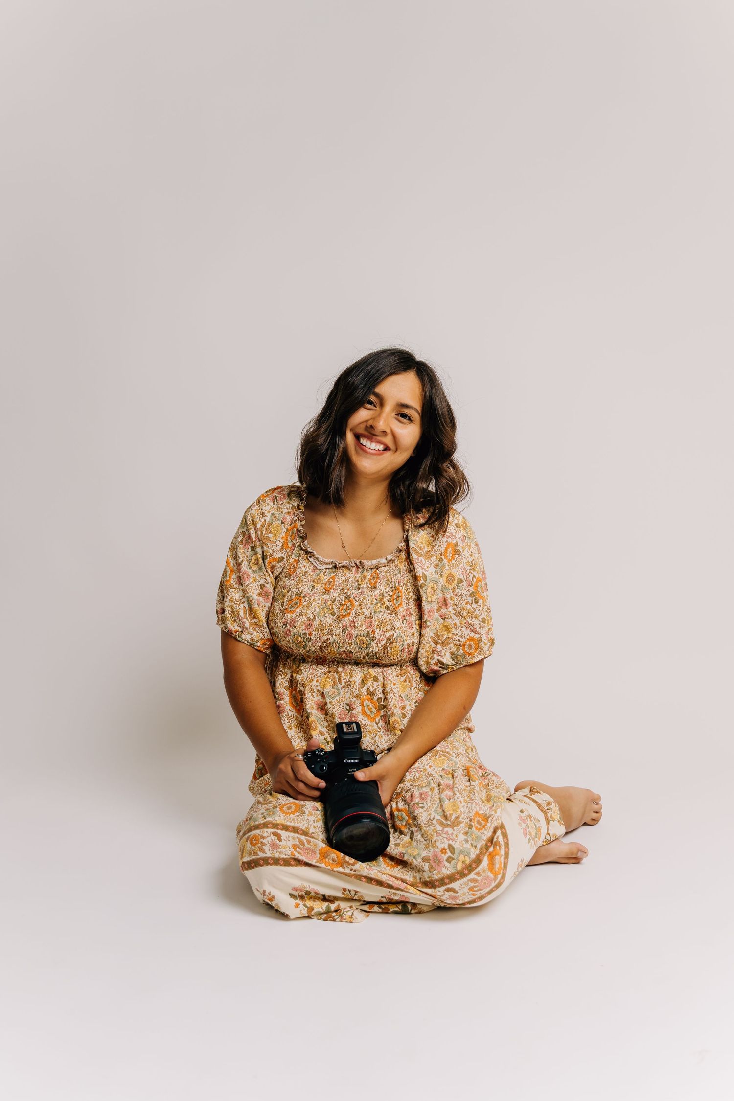 Smiling woman in floral dress sitting cross-legged on floor holding a camera against a light gray background.