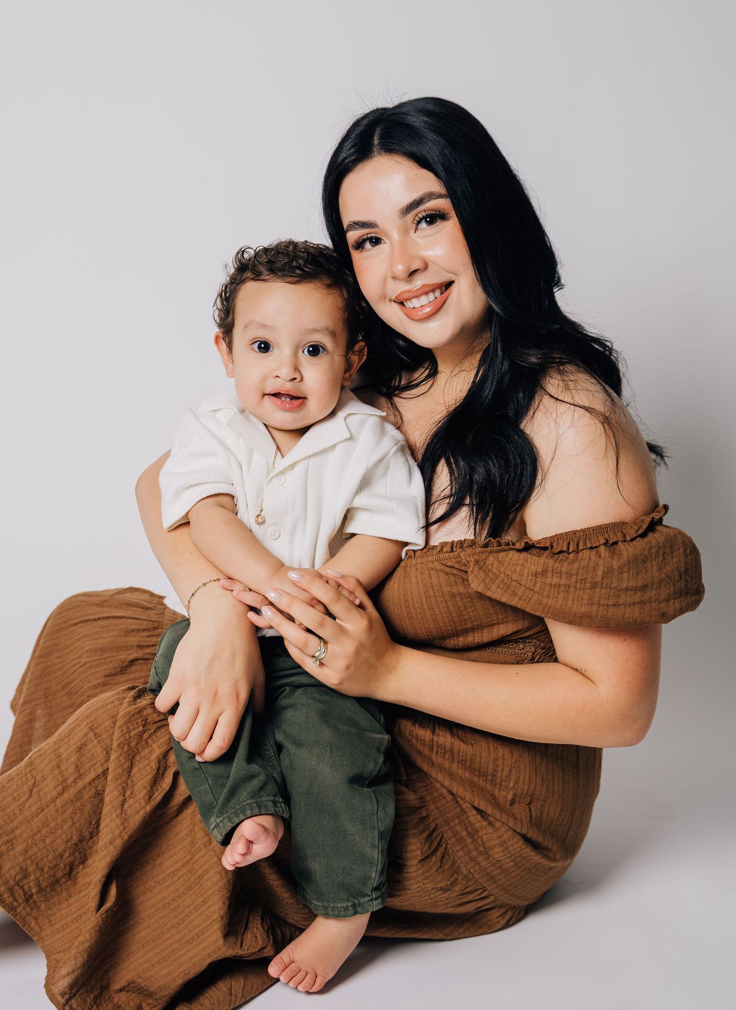 A smiling young woman in a brown off-shoulder dress sits holding a happy baby boy in a white shirt and green pants.