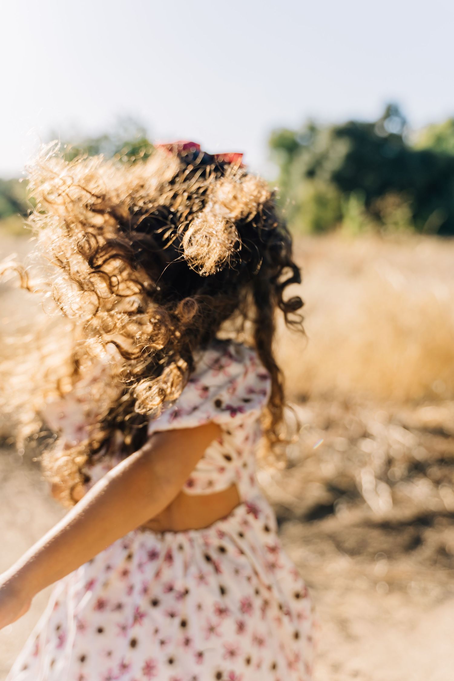 Curly blonde hair blows in the wind against a sunny outdoor backdrop with dried grass.