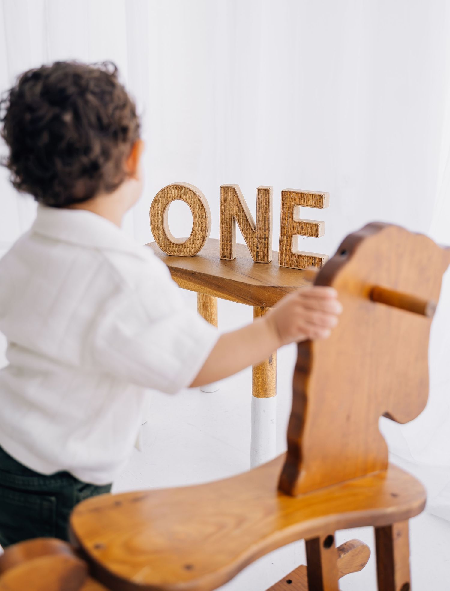 A toddler on a wooden rocking horse reaches for wooden ONE letters on a shelf for a first birthday photo.