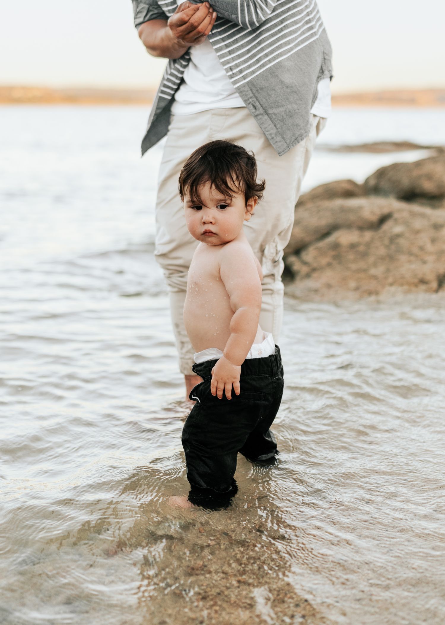 Small child stands in shallow water at the beach while being supervised.