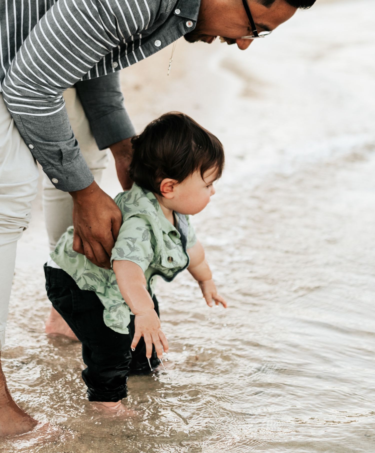 A gentle introduction to water play at the beach shore as adult supports small child testing the waves.
